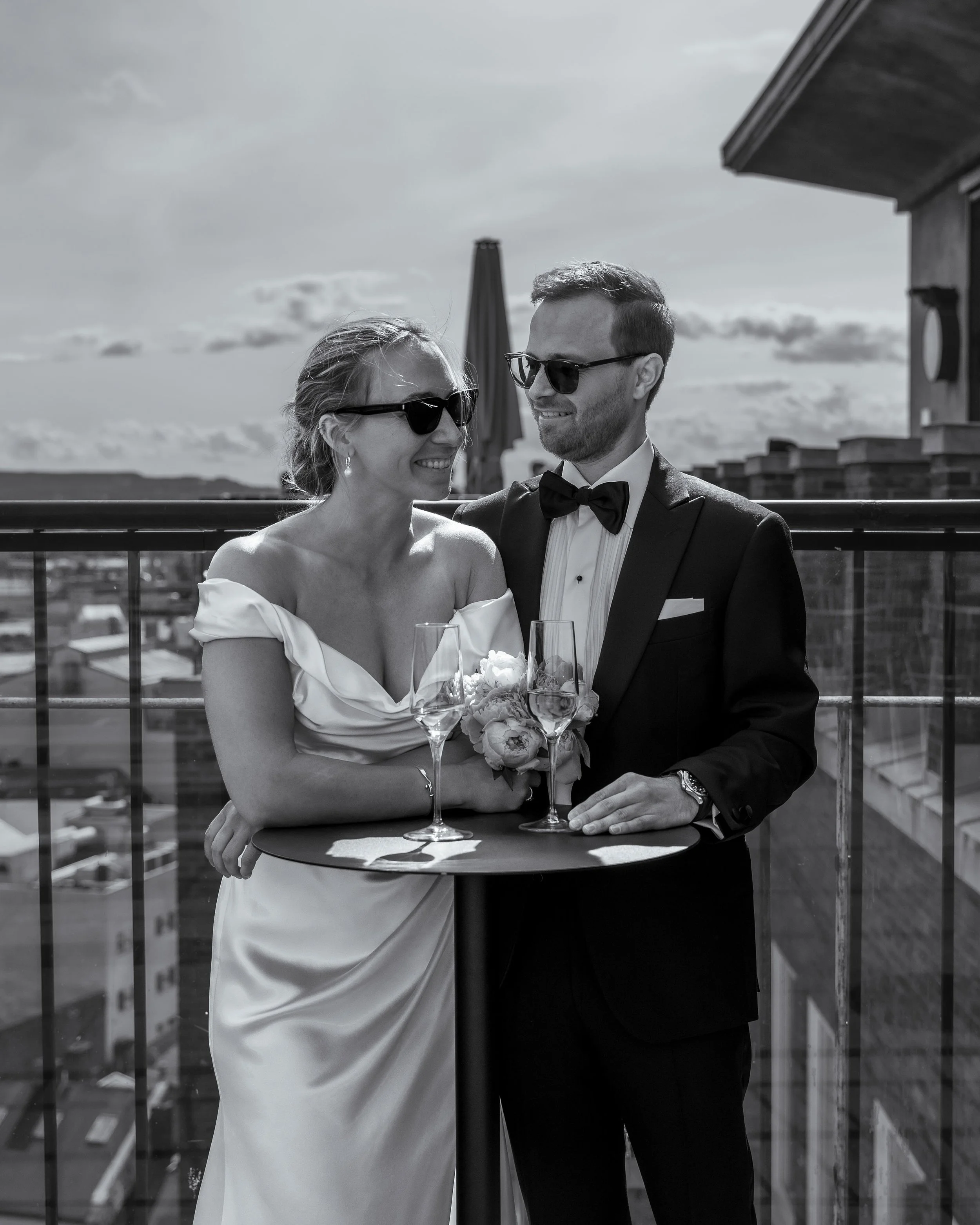 A black-and-white photo of a newlywed couple on a rooftop, smiling and looking at each other, with glasses of wine and a bouquet on a table in front of them, with city buildings and a cloudy sky in the background.