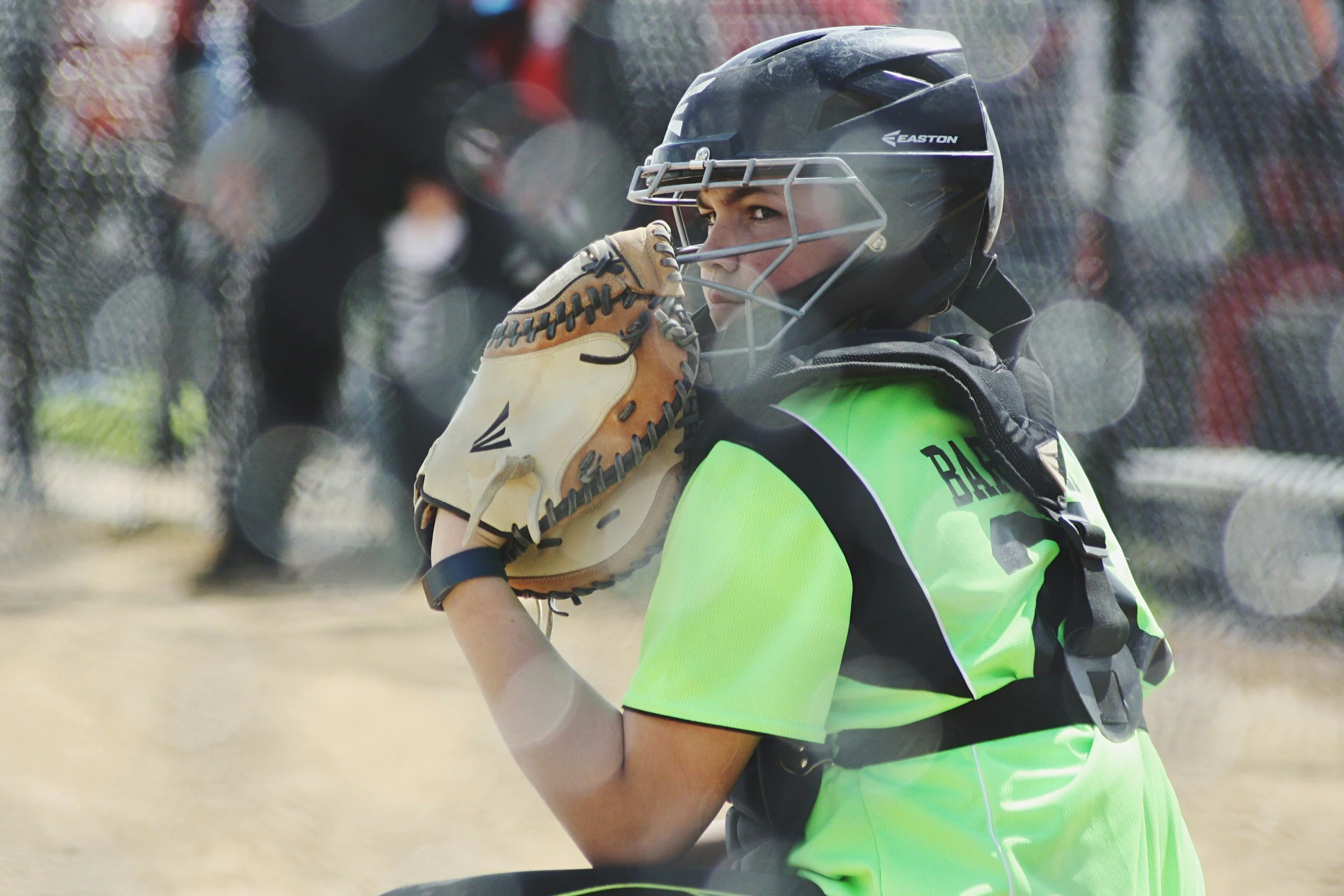 A baseball catcher wearing a black helmet with a face cage, a neon green jersey, and gloves, holding a baseball glove near his face at a baseball field.