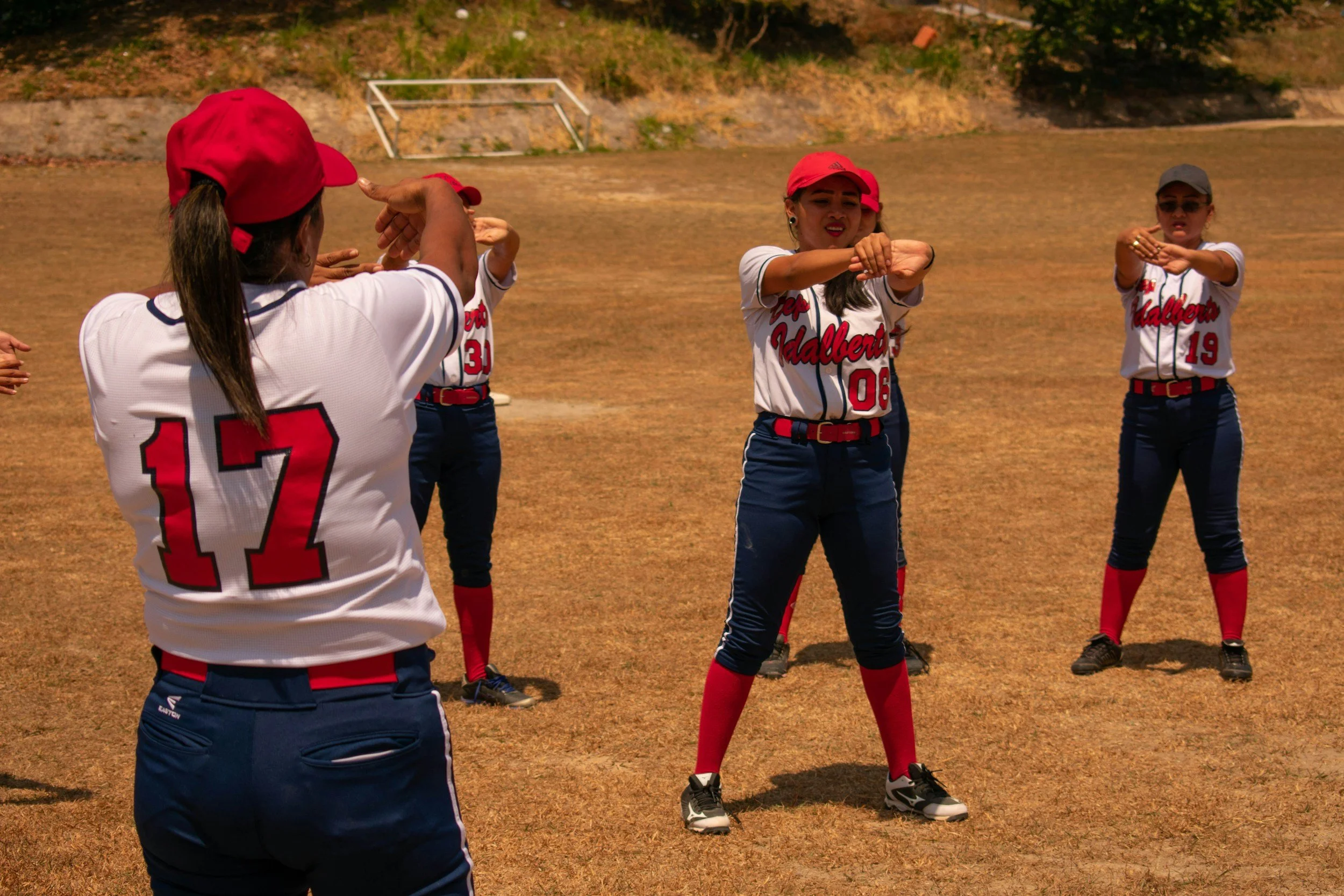 A group of women and girls practicing or warming up for a softball game on a field, wearing uniforms with red caps, white jerseys with red and blue accents, and dark pants. They appear to be stretching or doing warm-up exercises.