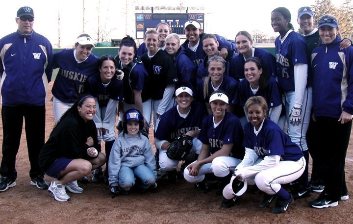 A group of female softball players and coaches posing on a softball field, wearing team uniforms and caps, smiling for a team photo.