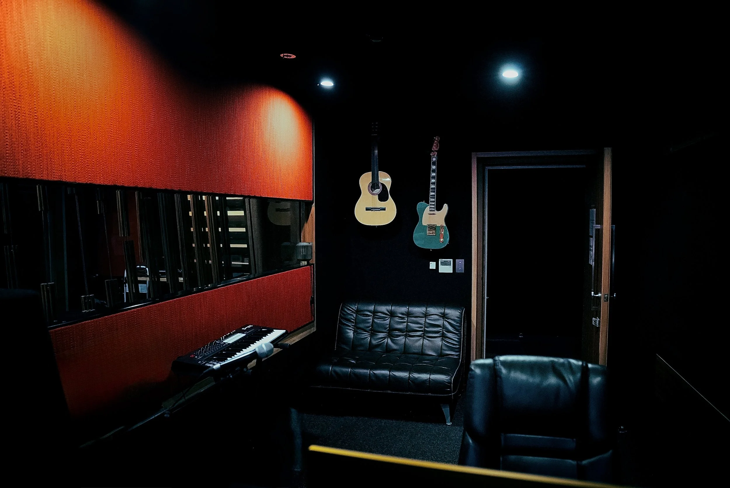 Music studio with mixing console, computer, speakers, and an individual sitting on a black leather chair in a room with red and black walls.