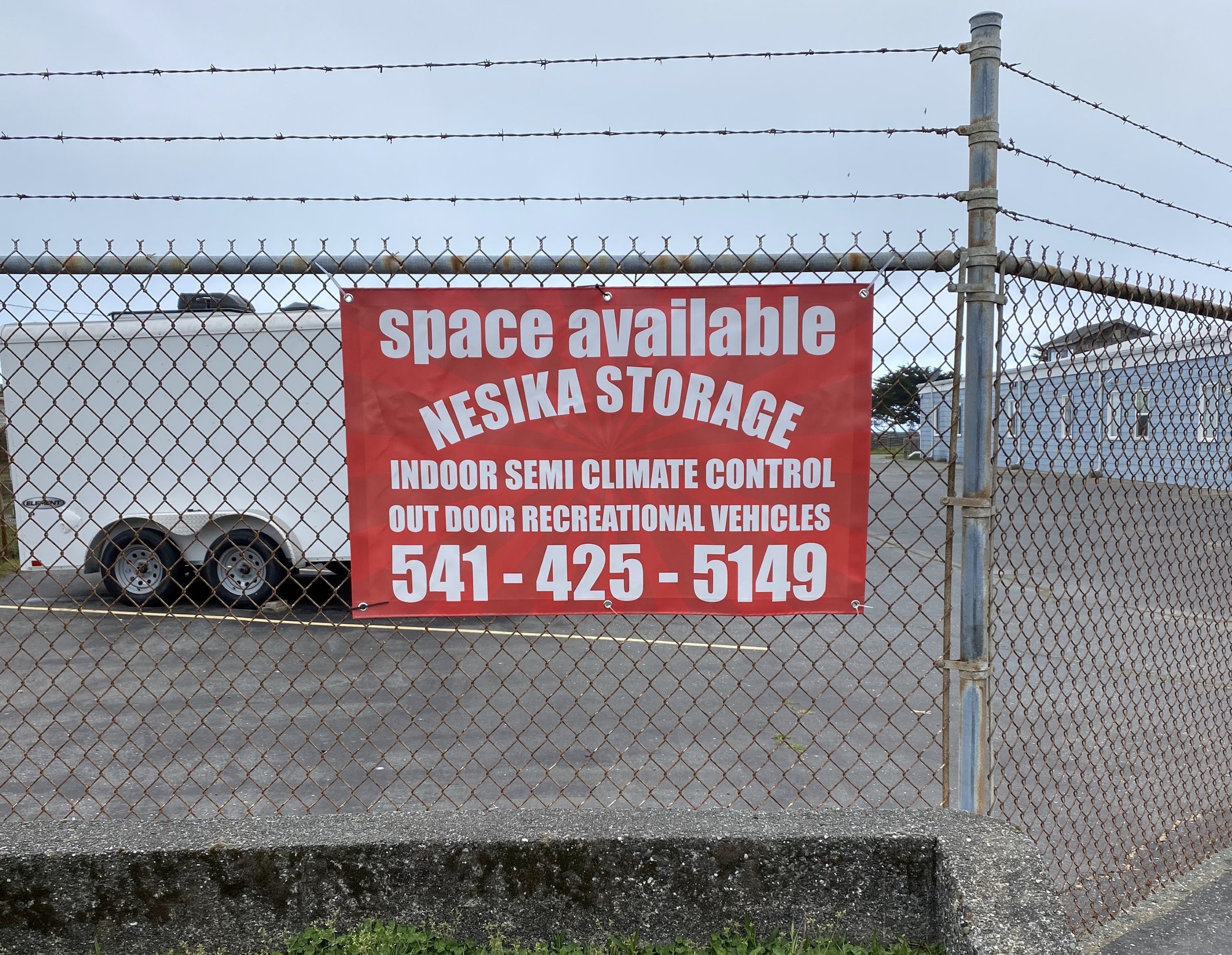 Red sign on a chain-link fence advertising space available at Nesika Storage with phone number 541-425-5149, mentioning indoor semi climate control and outdoor recreational vehicle storage.