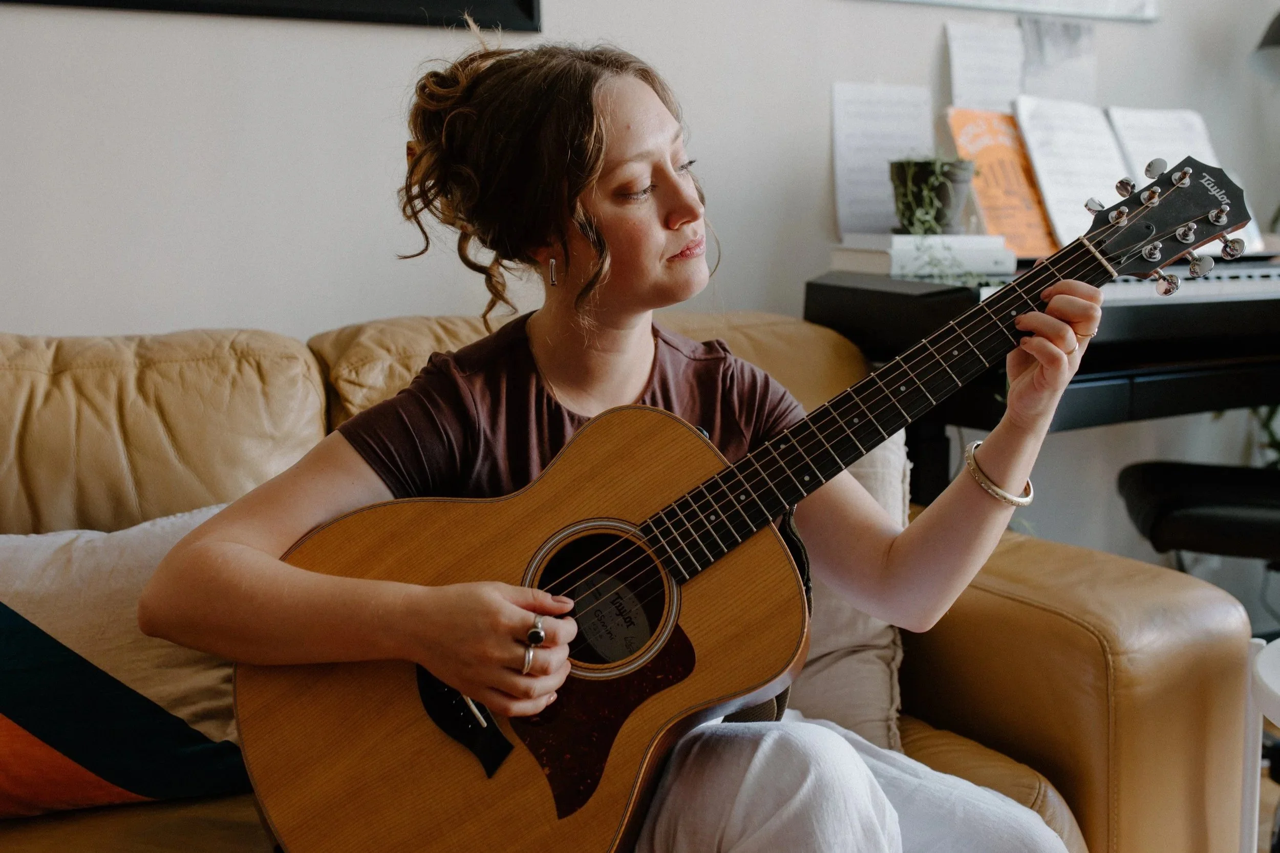 Olivia Apara sitting on her couch playing the acoustic guitar. She is a music therapist and teacher in Vancouver BC.