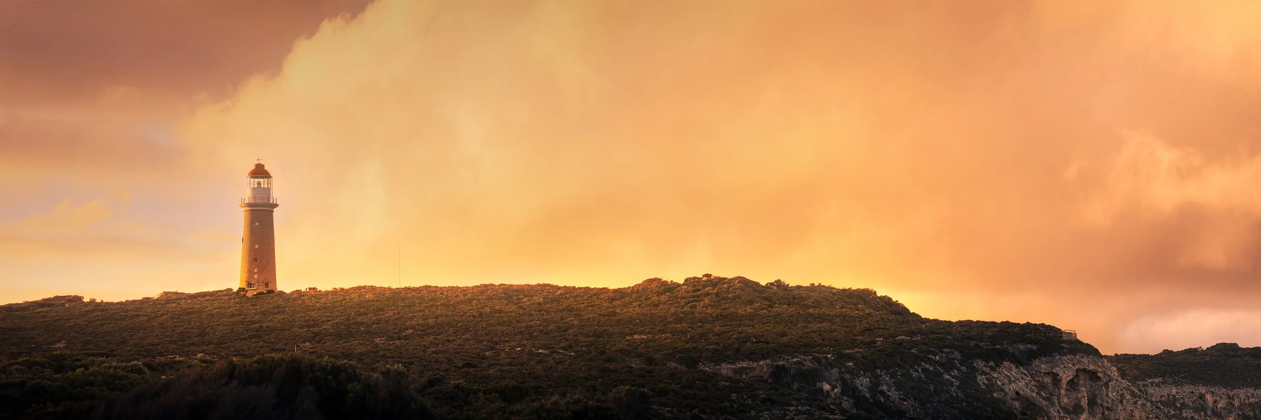 A lighthouse on top of a hill, sitting in front of a golden fluffy cloud bathed in sunset light.