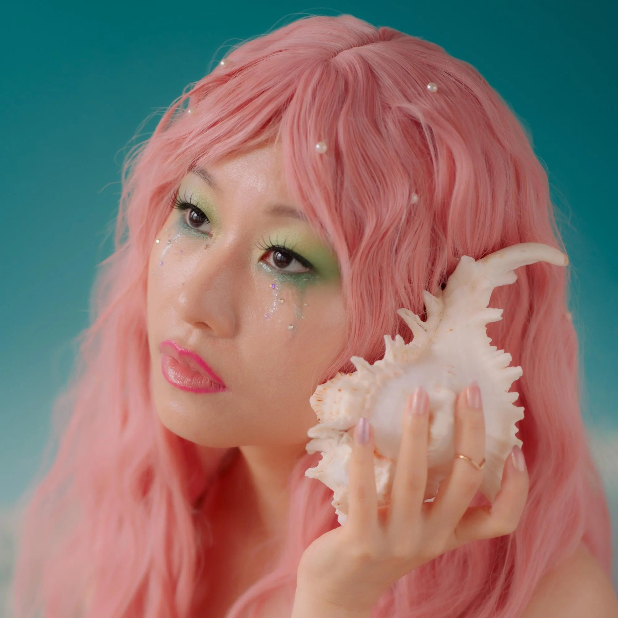 Sunny Daydream is an Asian femme with yellow-gold skin with wavy pink hair. They hold a conch seashell to their ear. Green eyeshadow runs down their face and pearls grow from their hair.