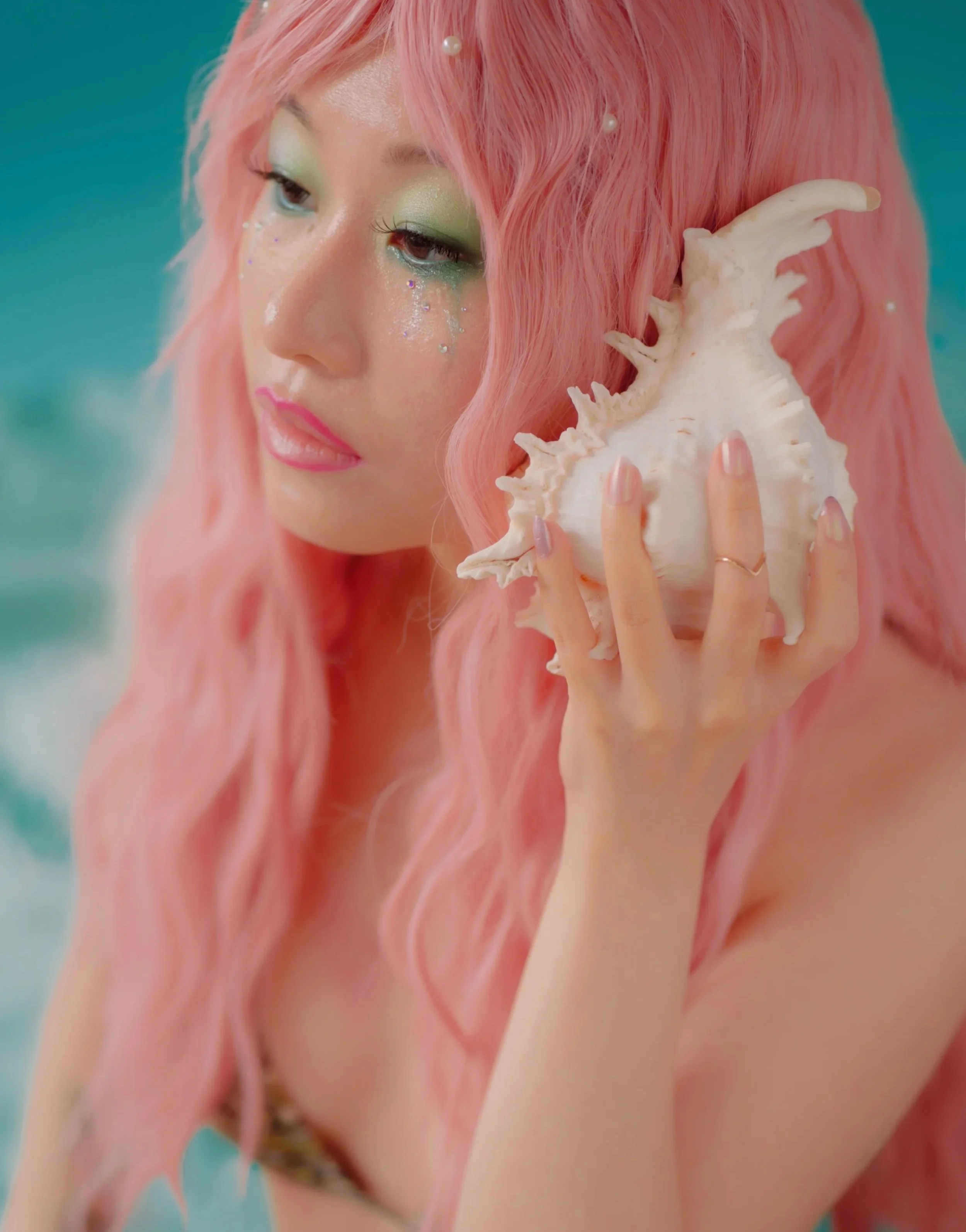 An Asian femme with long pink hair holds a large conch seashell to their ear with green eyeshadow running down their face. Pearls grow out of their hair and their nails are painted a dusty pink. They wear a single gold ring on their third finger.
