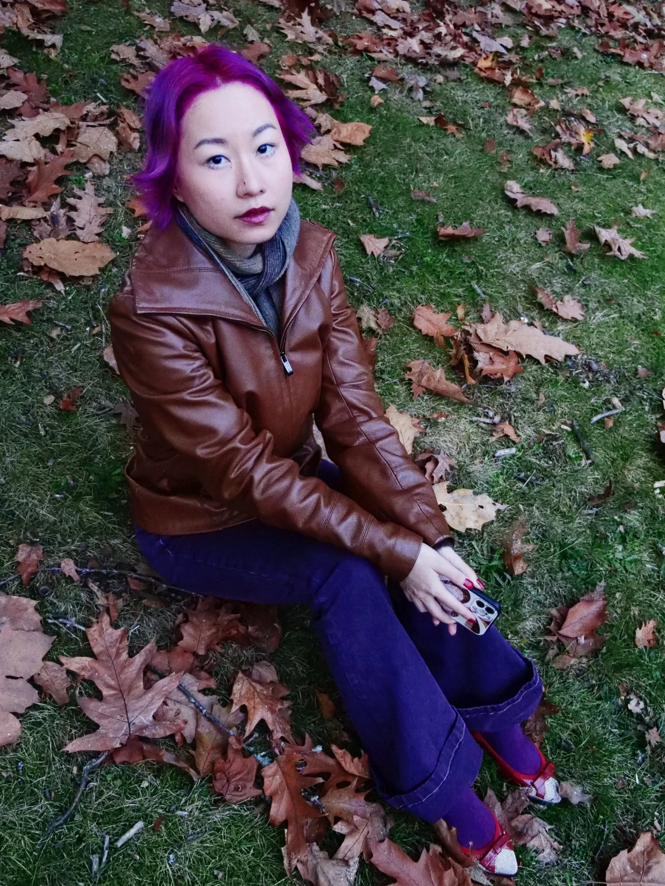A feminine young person with purple hair sits on grass, surrounded by fallen leaves.