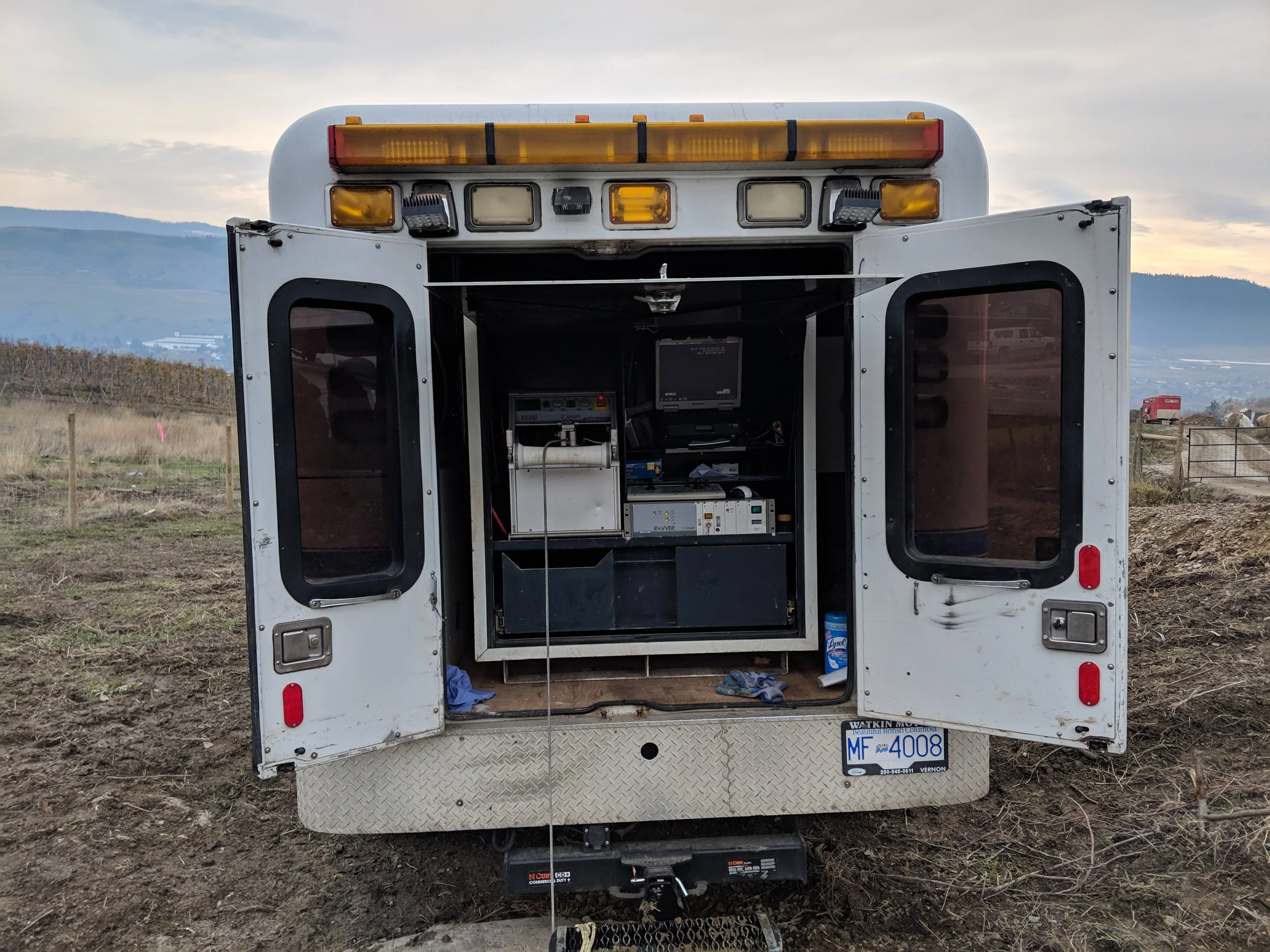 Mobile medical or emergency vehicle with the back doors open, showing medical and electronic equipment inside, parked outdoors in a rural area with hills in the background.