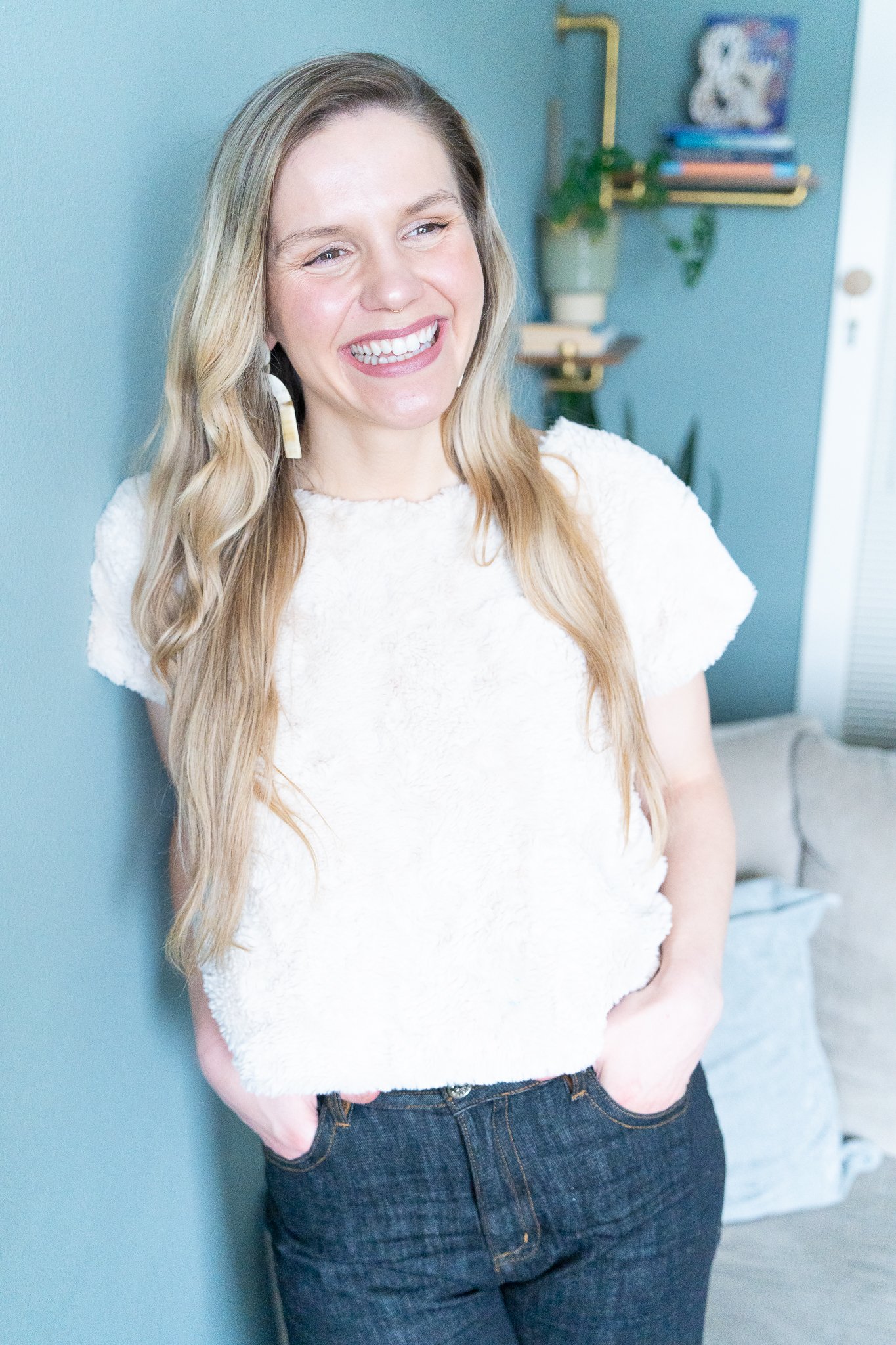 A woman with long blonde hair, wearing a white textured top, smiling with her hands in her pockets, standing indoors against a teal wall with decorative shelves and plants.