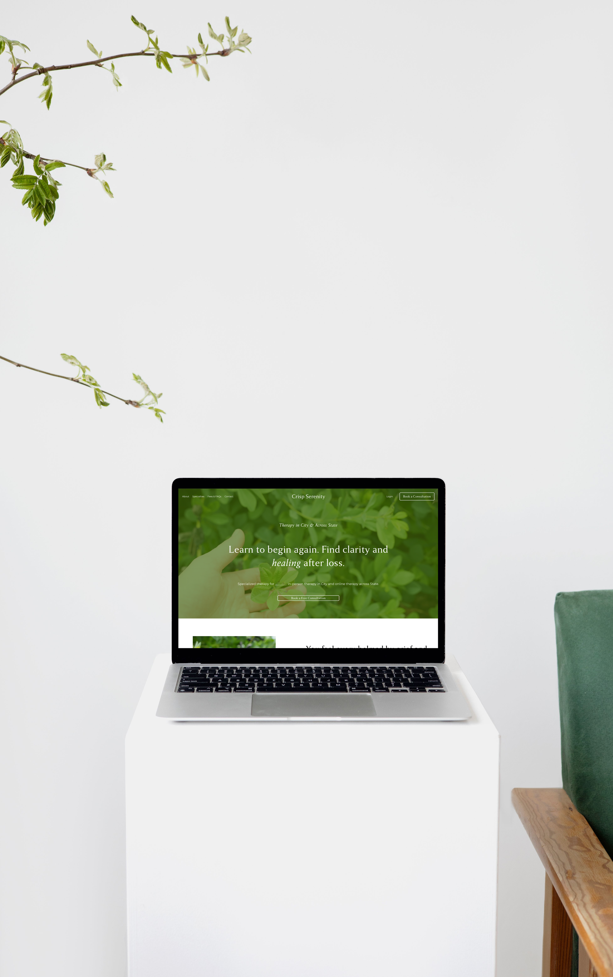 A laptop on a white table displaying a website with green and white colors, surrounded by a wall with a branch and leaves on the upper left and a wooden table with a green cushioned chair on the right.