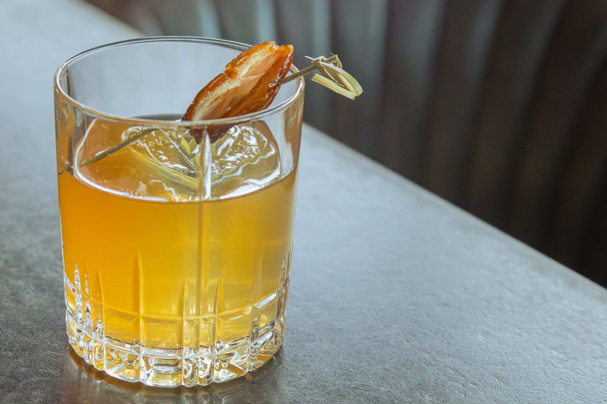 A glass of whiskey garnished with a strip of bacon on a toothpick, with ice cubes in the drink.