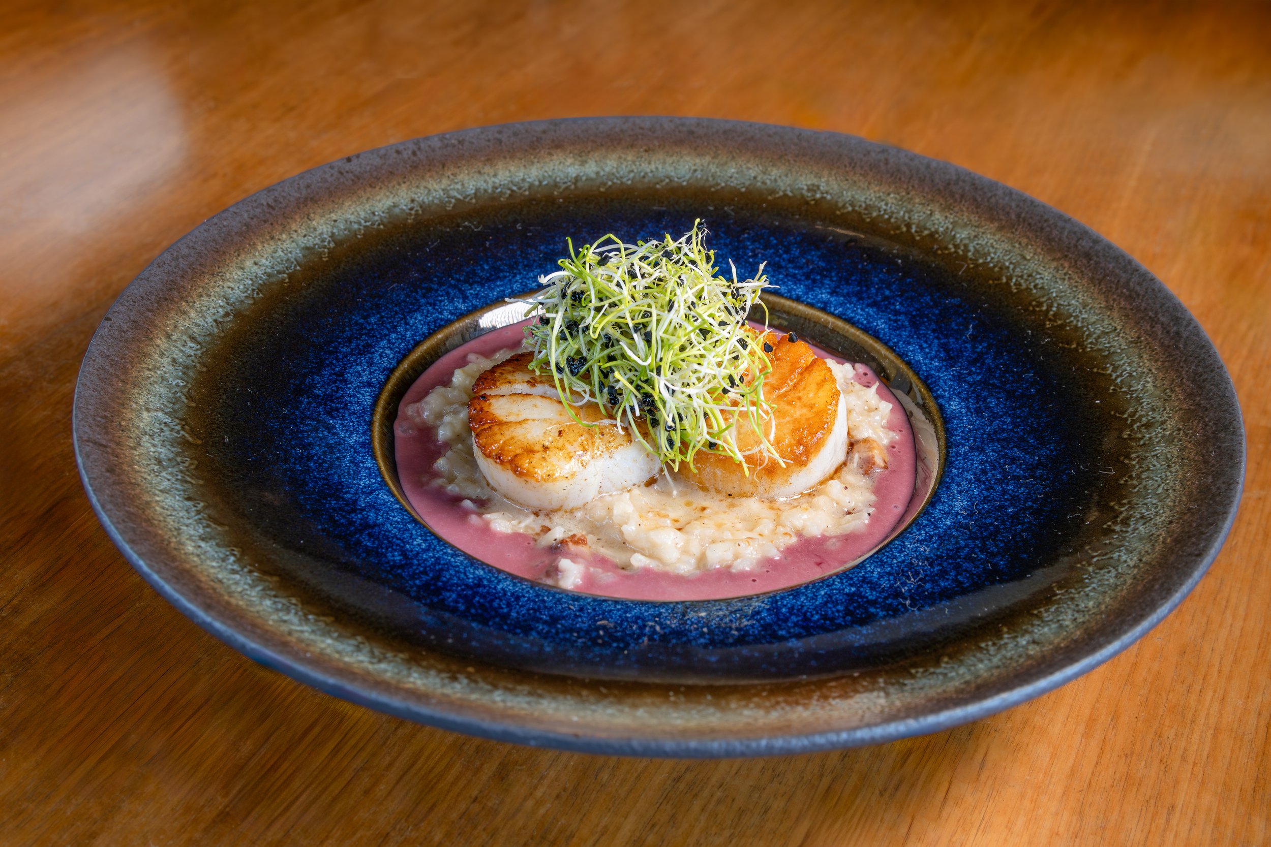 Seared scallops garnished with microgreens on a pink sauce, served on a textured black and blue plate.