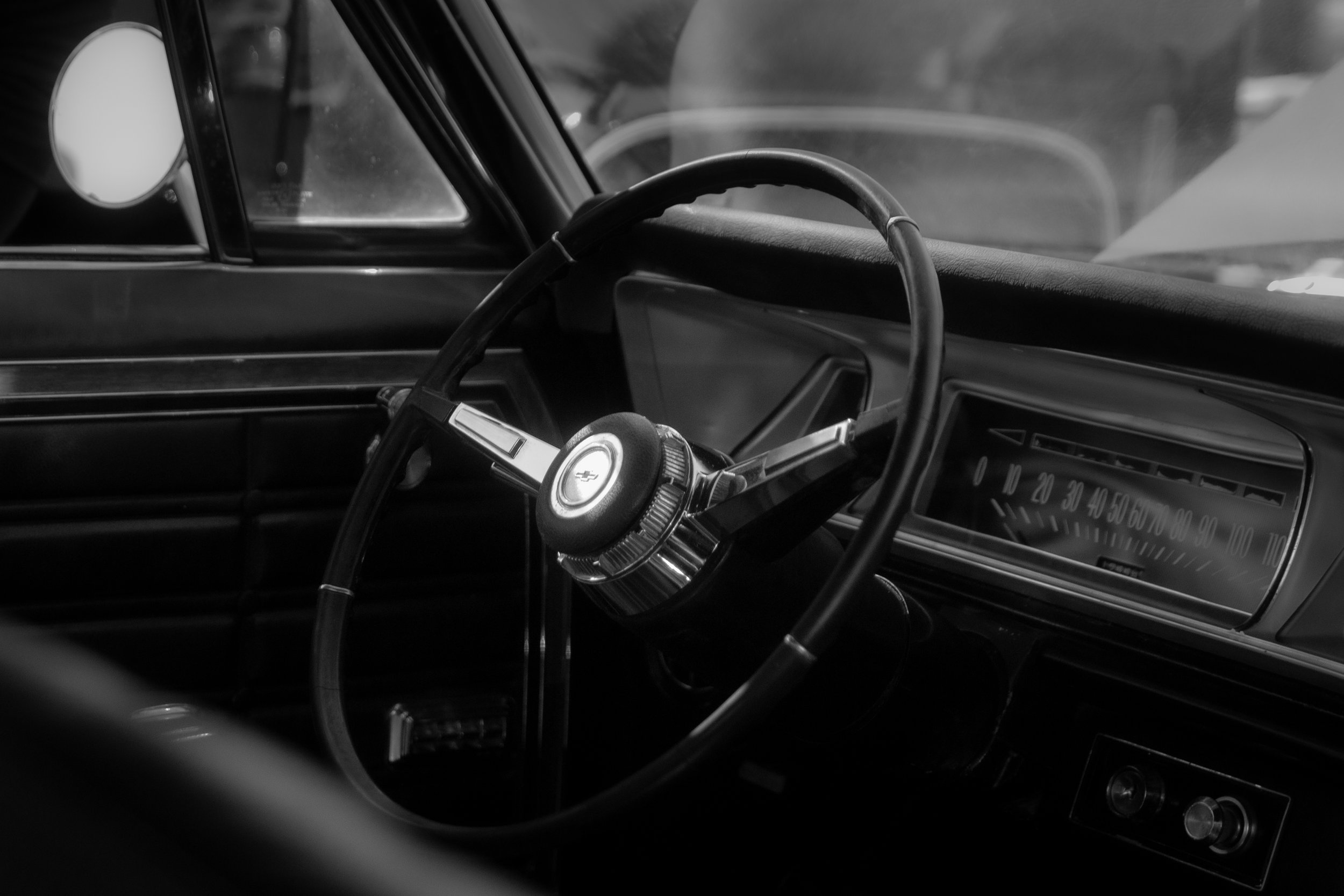 Black and white photo of the interior of a vintage car, showing the steering wheel, dashboard, and speedometer.