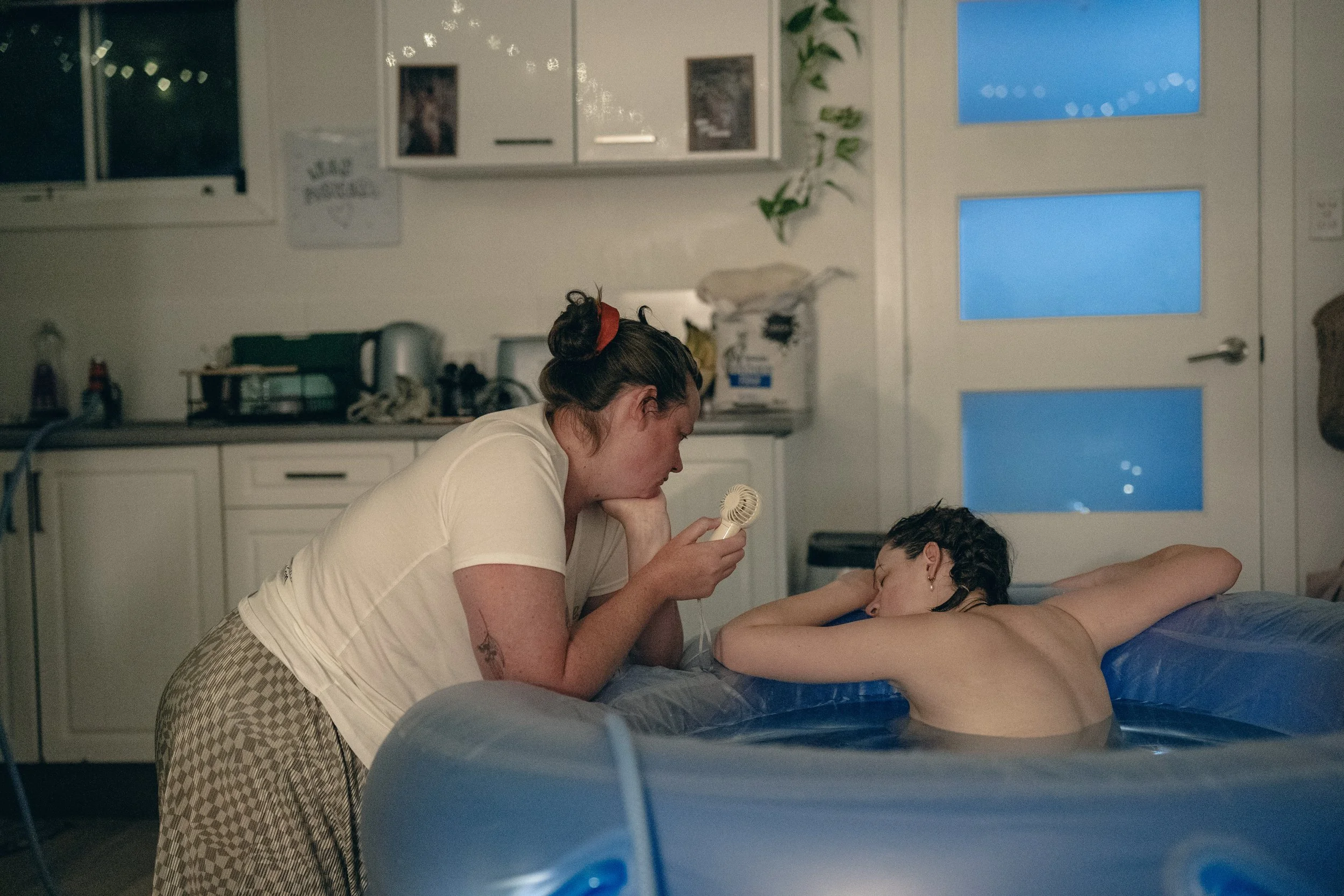 Two people in a dimly lit room, one sitting in an inflatable pool, and the other leaning over with a fan, providing support.