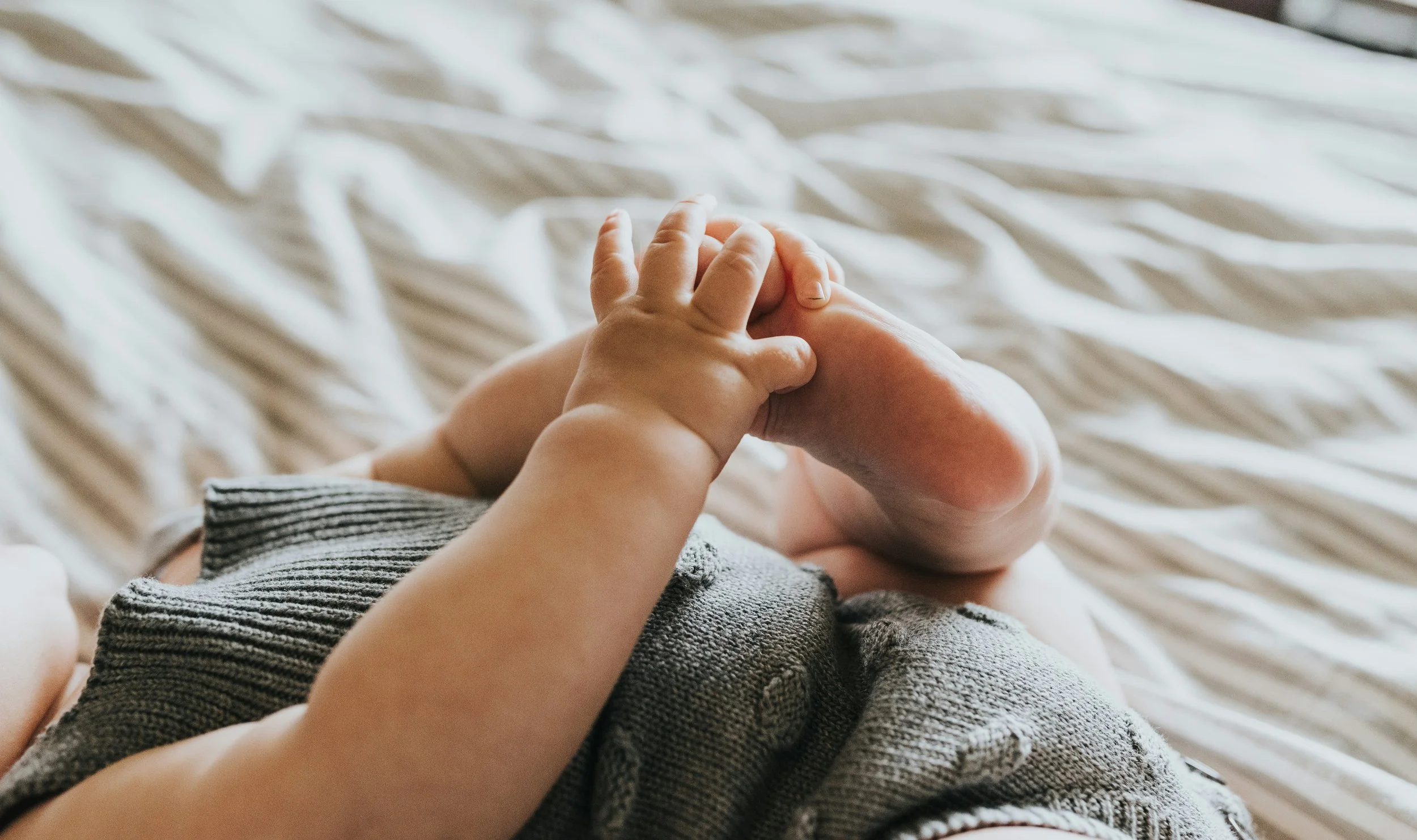Close-up of a baby lying down, holding its foot with both hands, wearing a knitted outfit.