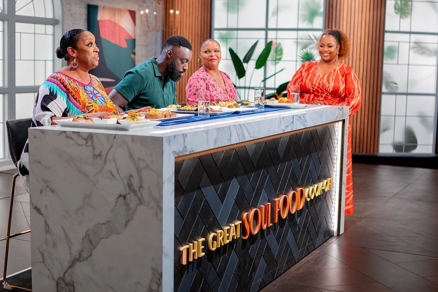 Four women sit behind a marble-topped counter with plates of food in front of them at an event or food tasting. The counter has a sign that reads "The Great Soul Food Cook". The background features large windows, wooden decor, potted plants, and abstract art.