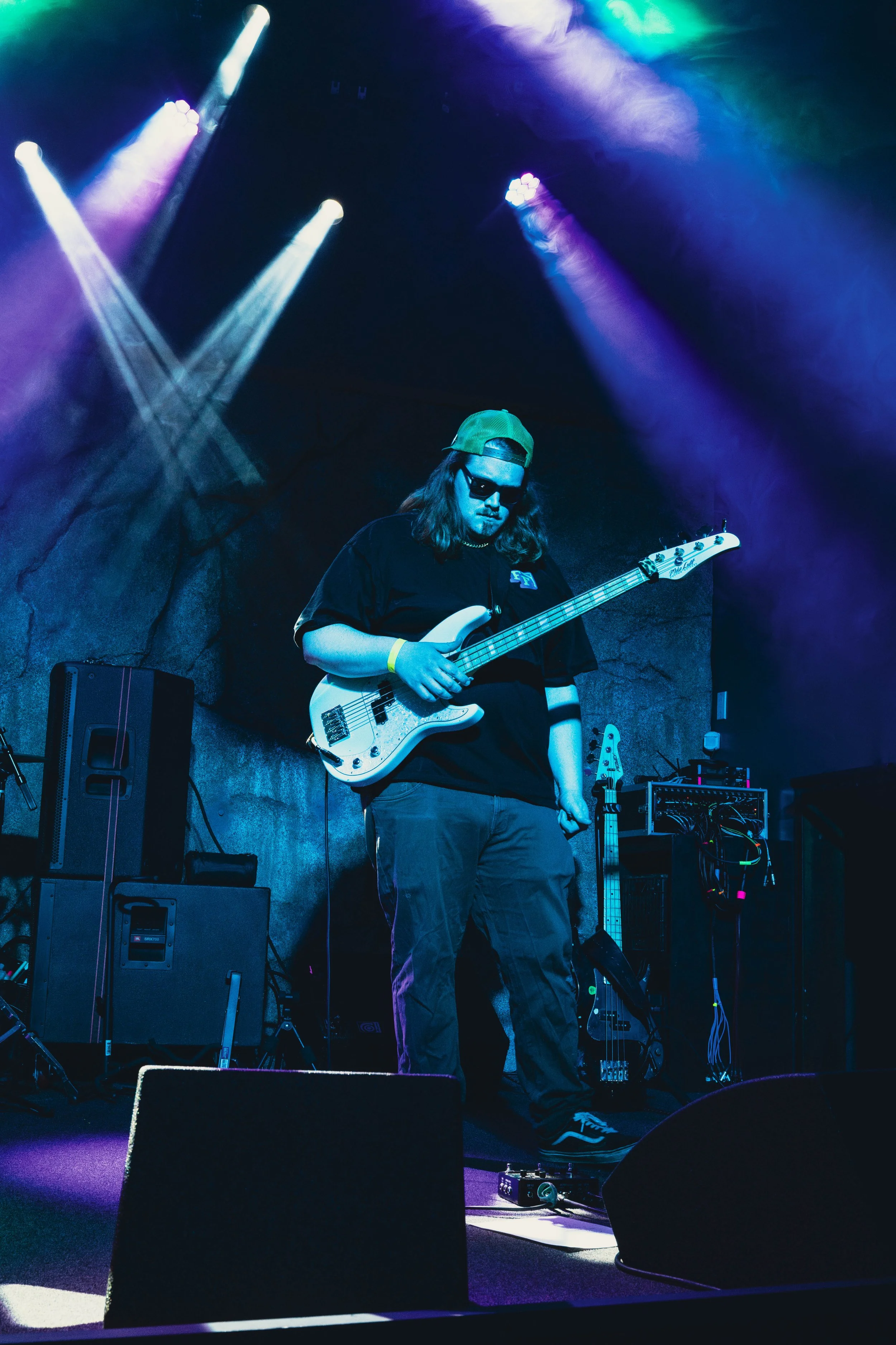 A musician with long hair, sunglasses, a baseball cap backwards, and a black t-shirt, playing an electric bass guitar on stage under purple, blue, and green stage lights.