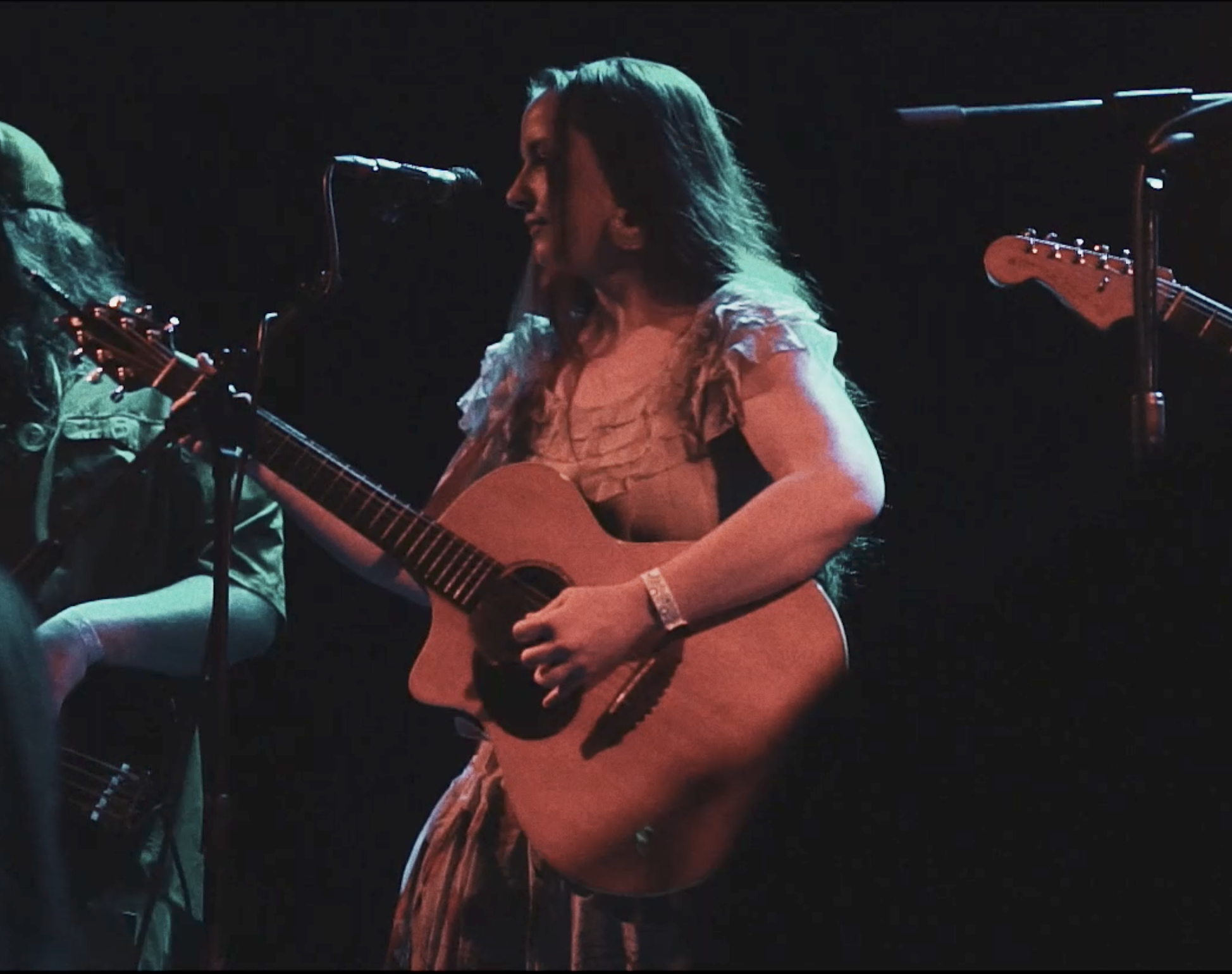 A woman with long hair playing an acoustic guitar on stage, singing into a microphone, with another musician partially visible on the side.