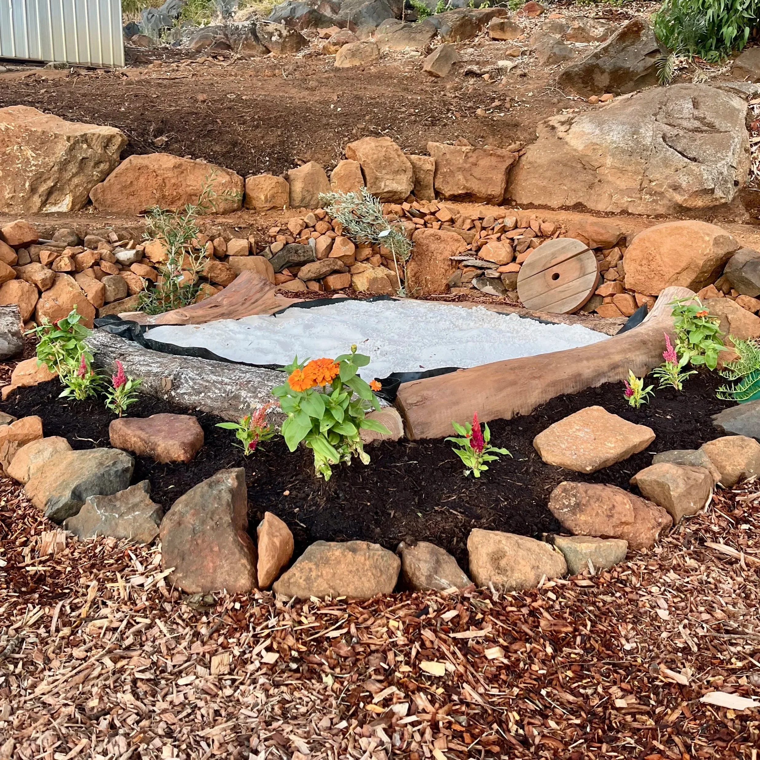A garden with a small sandbox surrounded by rocks, soil, and blooming flowers, with a natural log lining.