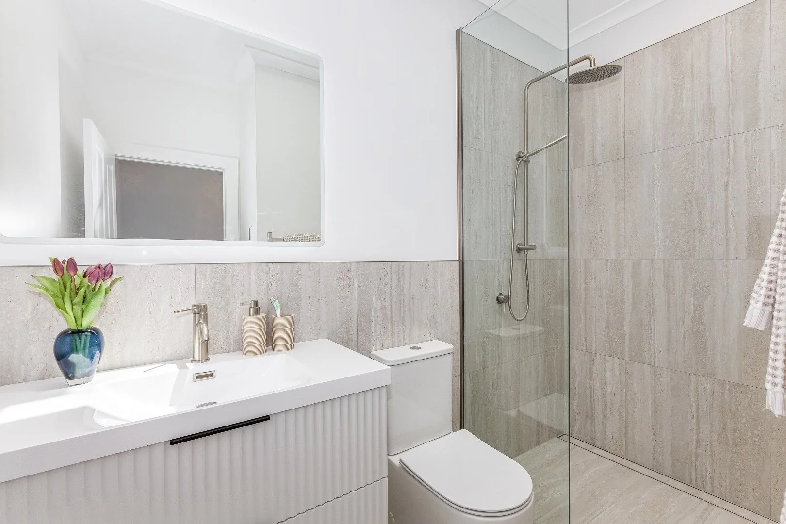 Modern bathroom with a white vanity, toothbrush holder, soap dispenser, a toilet, a large mirror, and a walk-in shower with glass door and rain shower head.