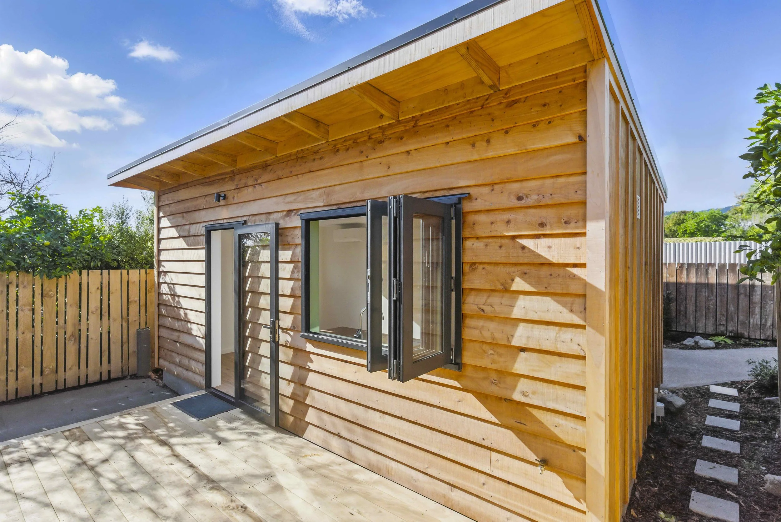 Small wooden house with open black-framed window and glass door, outdoors with wooden fence, trees, and blue sky.