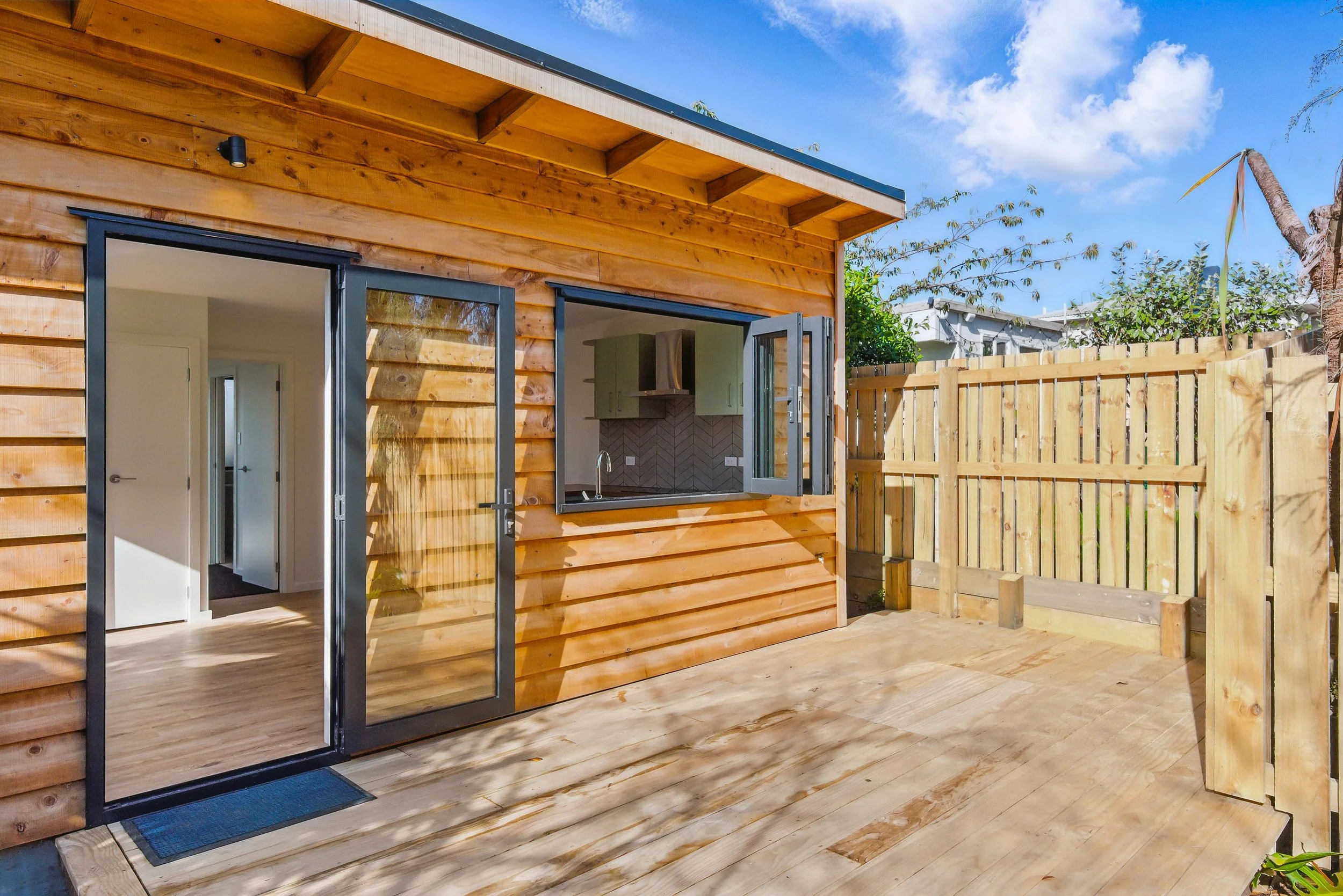 A small backyard patio with wooden deck, wooden fence, and open door leading into a house with a kitchen visible through a window.