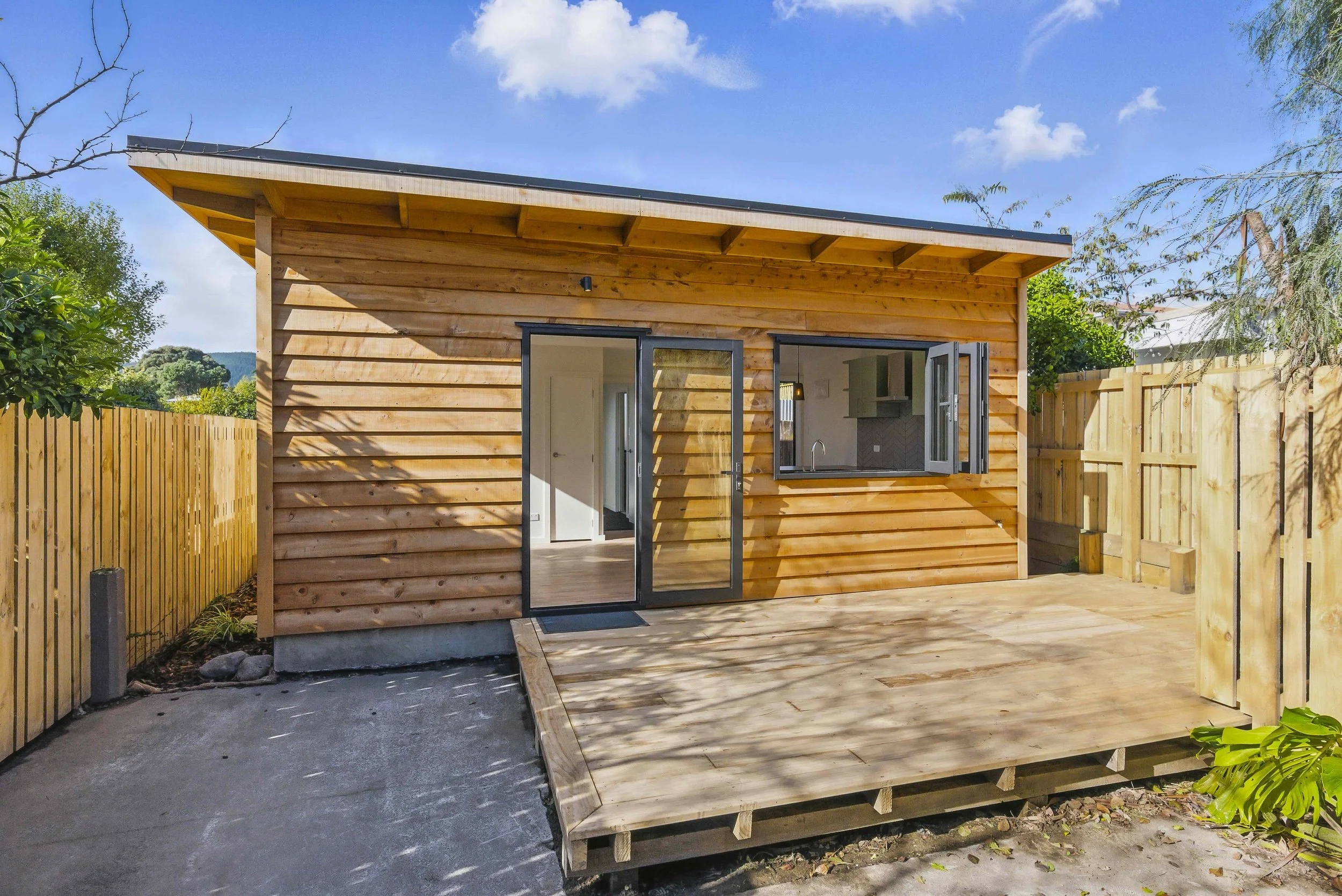 Small wooden house with an open door and window, wooden deck, surrounded by a wooden fence, on a sunny day with blue sky.