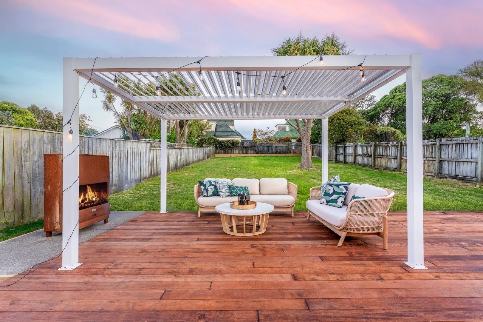Outdoor patio with wooden deck, white pergola, string lights, beige and wicker sofas with patterned throw pillows, a round coffee table, fire pit, picnic table, green lawn, trees, and house in background at sunset.