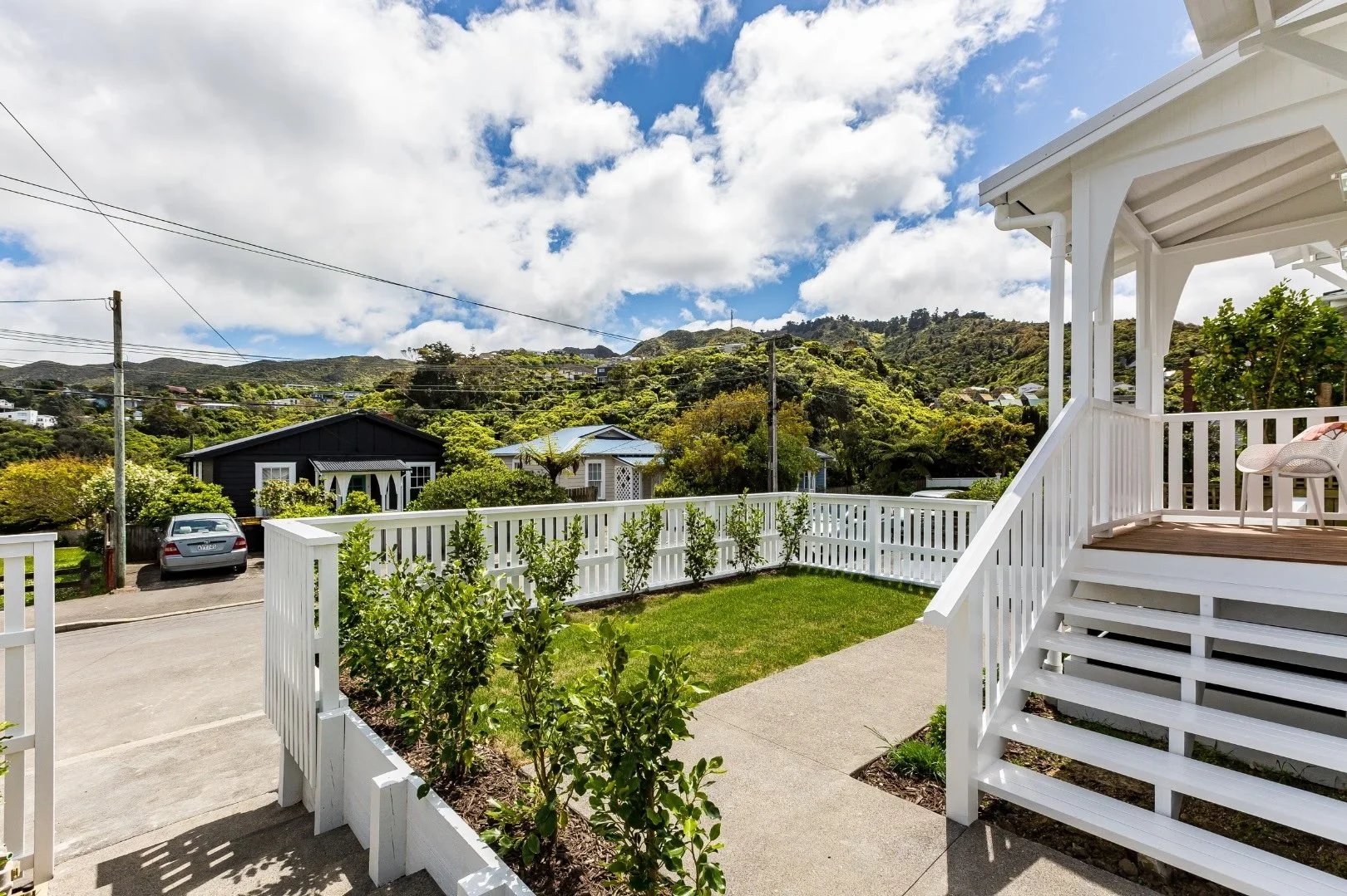 A view from a porch showing a grassy backyard, a white fence, and a scenic hillside with houses and trees under a partly cloudy sky.