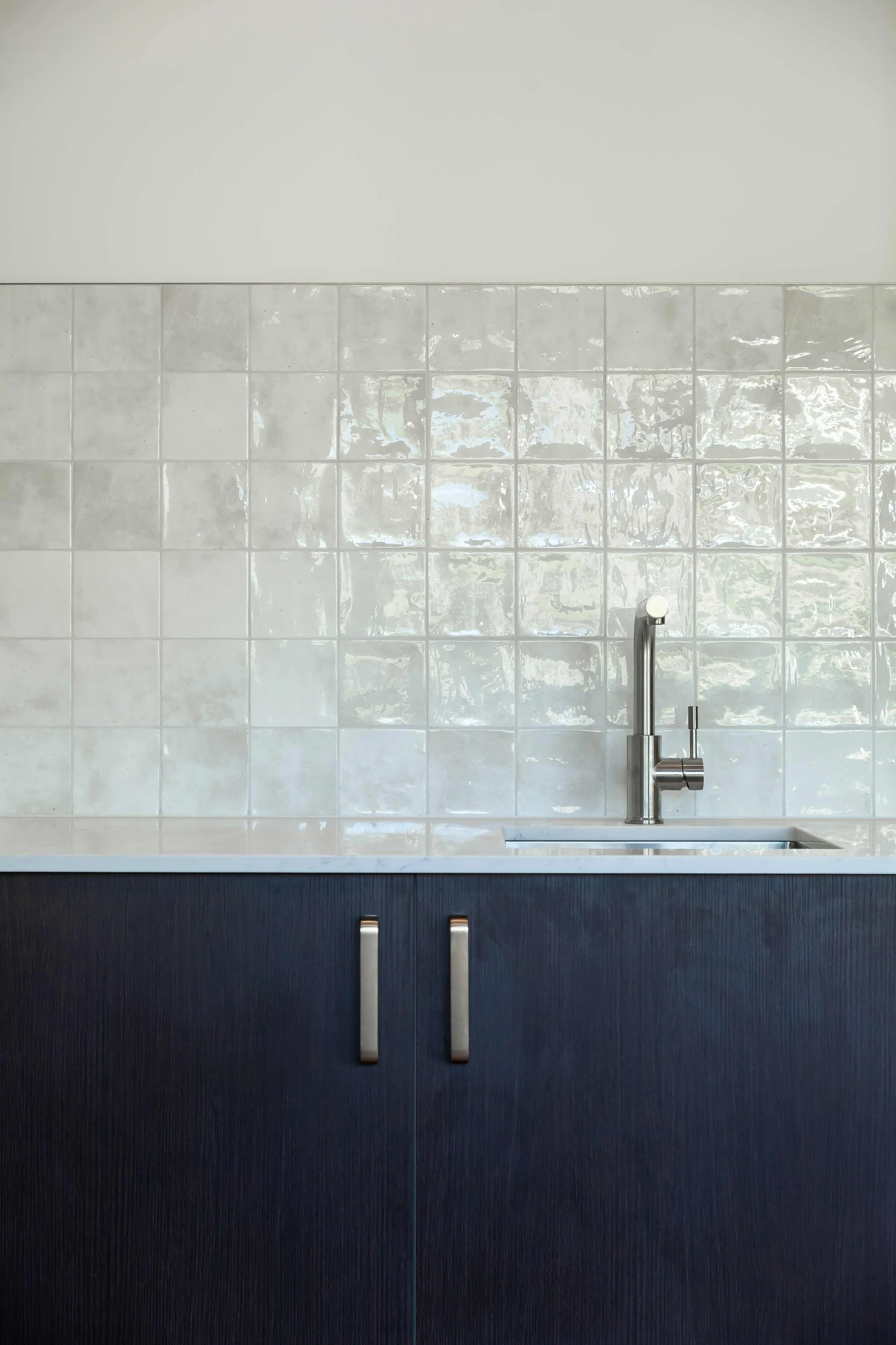 Kitchen with a dark blue cabinet, a white countertop, and a stainless steel faucet over a sink, white tiled backsplash, and a plain white wall above.