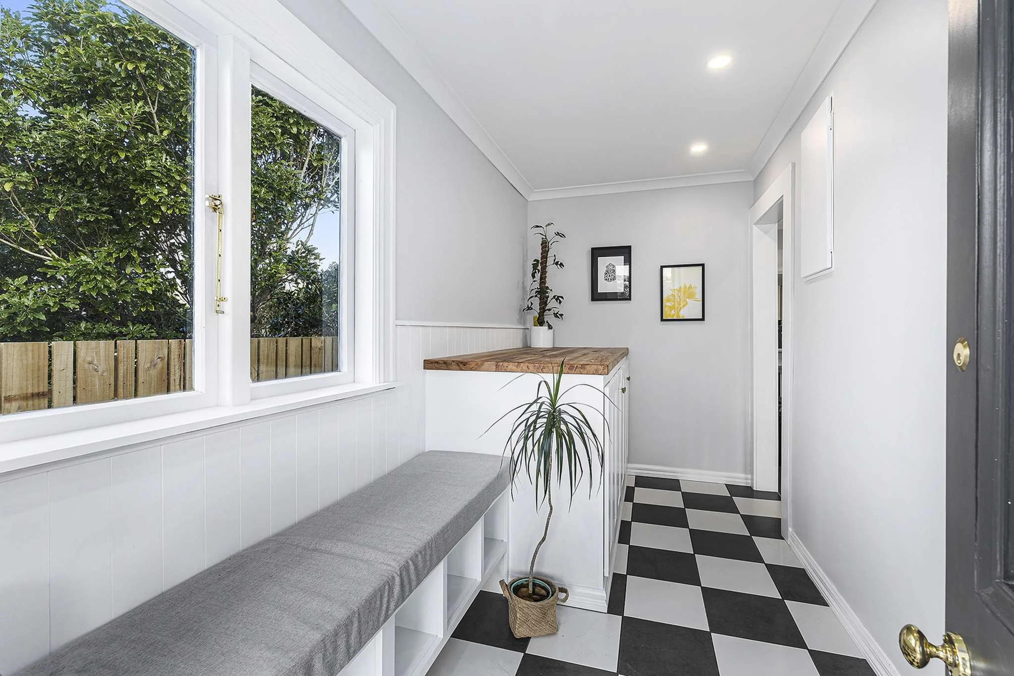 An indoor space with white walls, a black and white checkered floor, a window showing green foliage outside, a gray bench with cubbies, a potted plant, framed artwork, and a wooden countertop.