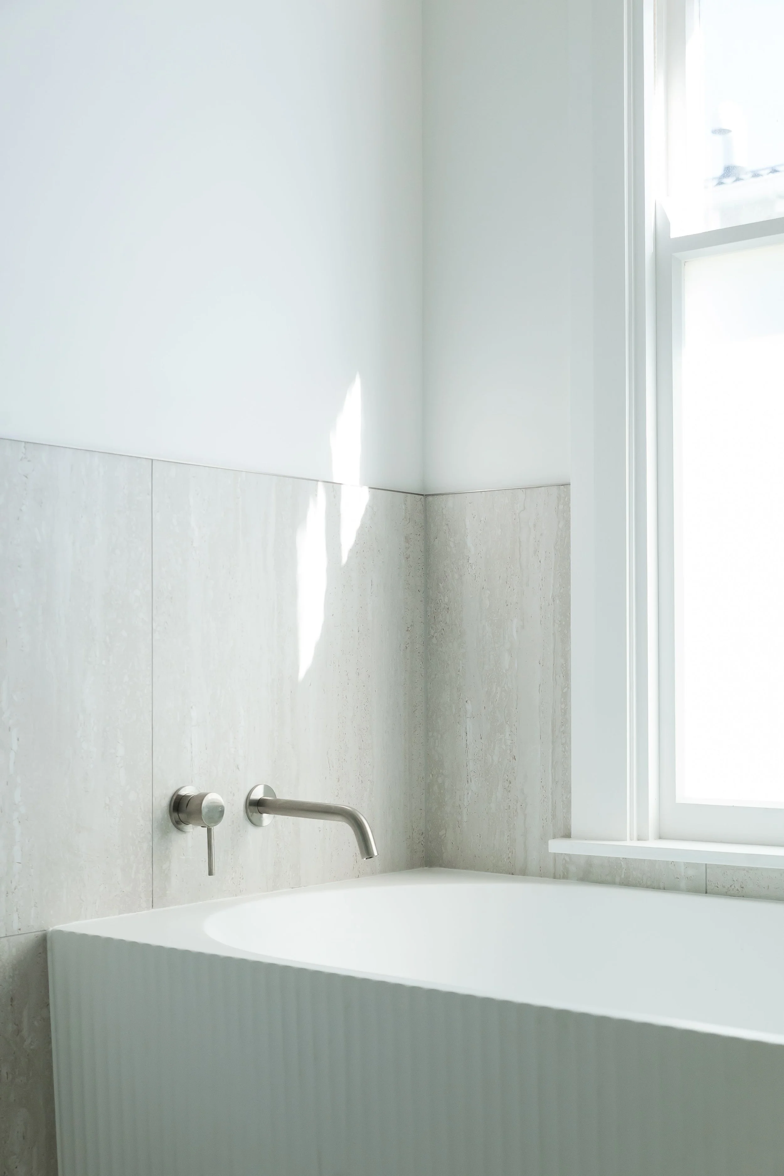 White bathtub in a bright, minimal bathroom with a large window, concrete wall tiles, and a wall-mounted faucet.