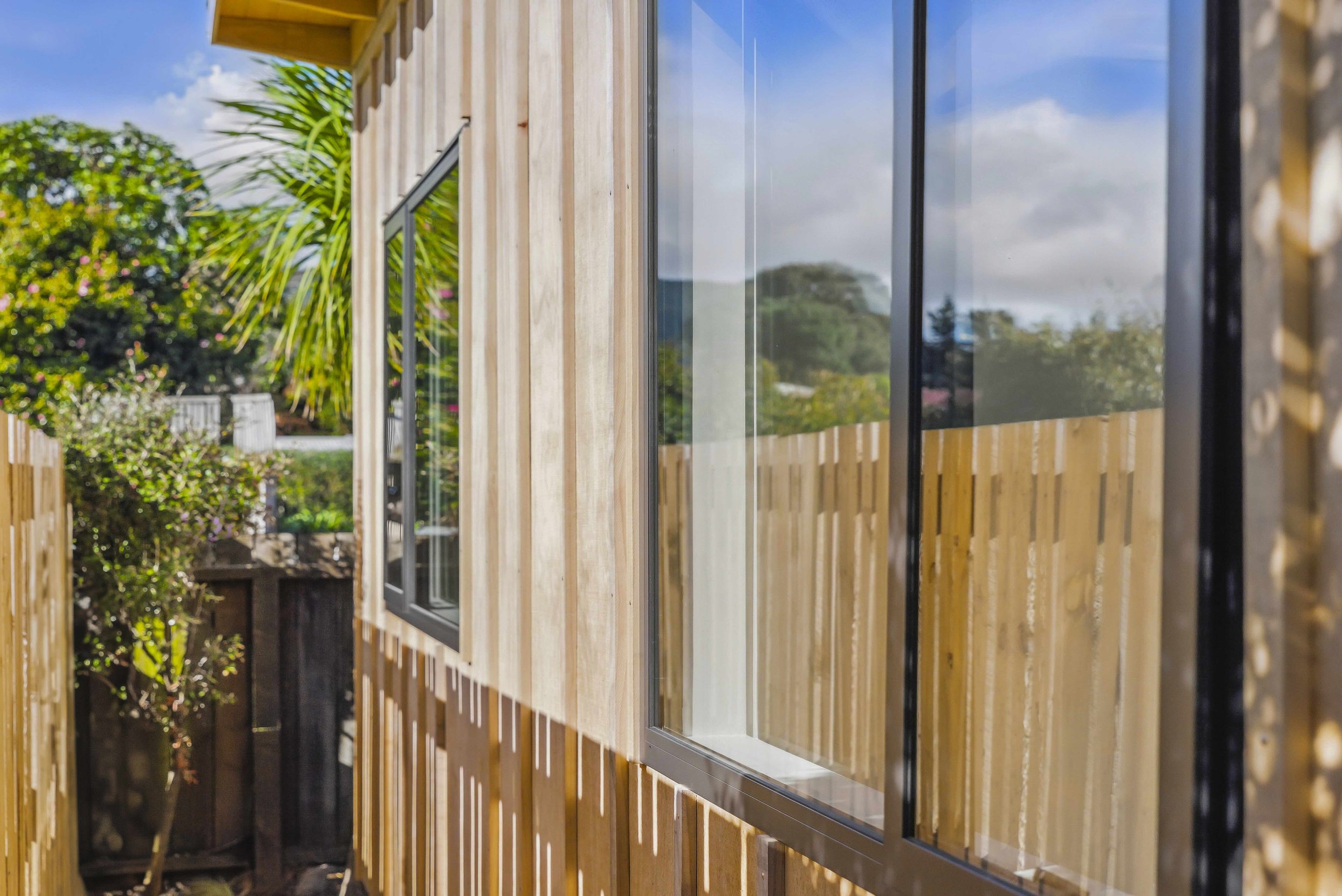 Close-up of a wooden exterior wall of a building with large glass windows, with greenery and trees in the background under a partly cloudy sky.