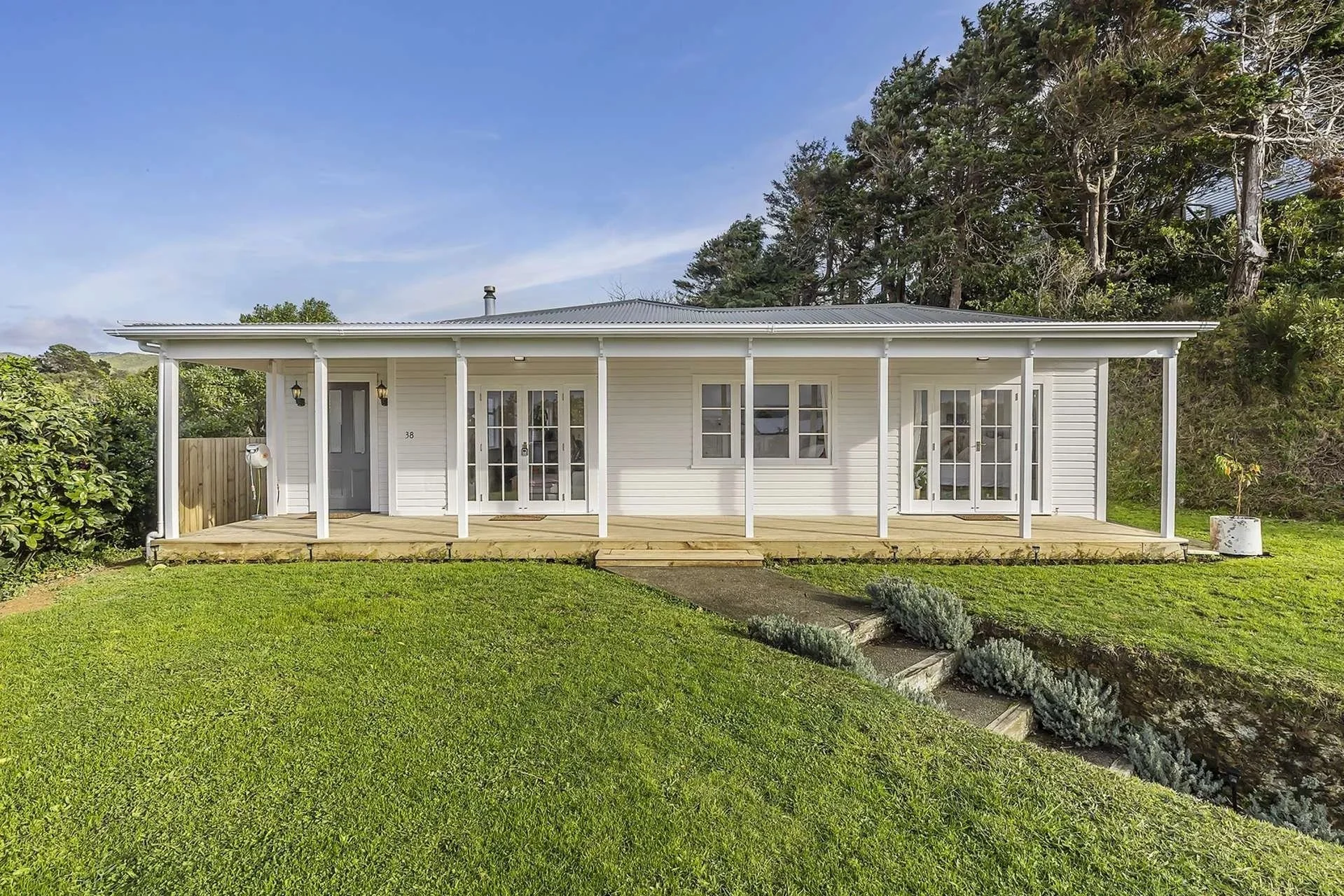 White house with a wooden porch, multiple glass-paneled doors and windows, surrounded by a well-maintained lawn and trees in the background, under a clear sky.