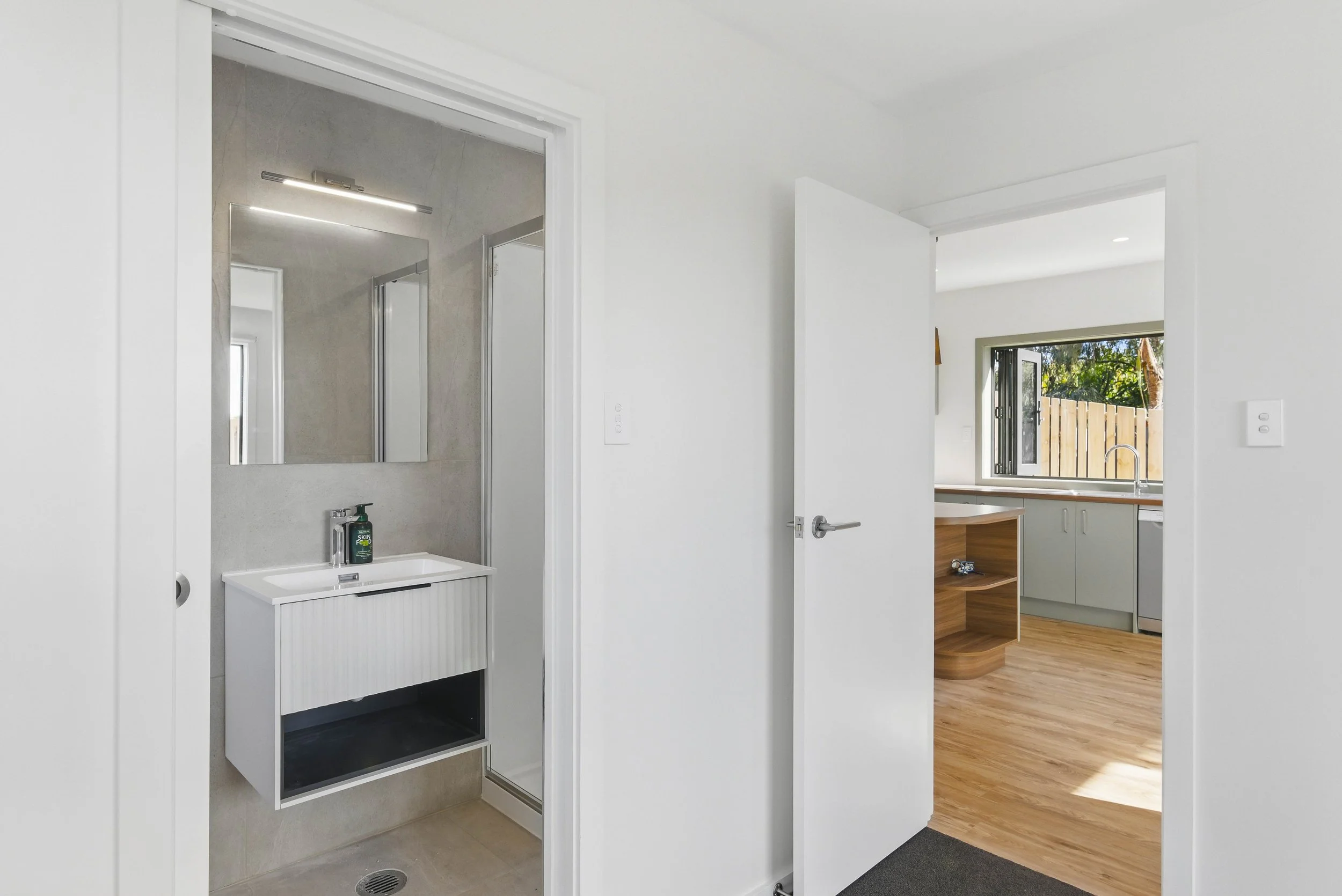 View of a bathroom with a white sink vanity and a mirror, open door leading to a kitchen with a window and wooden flooring.