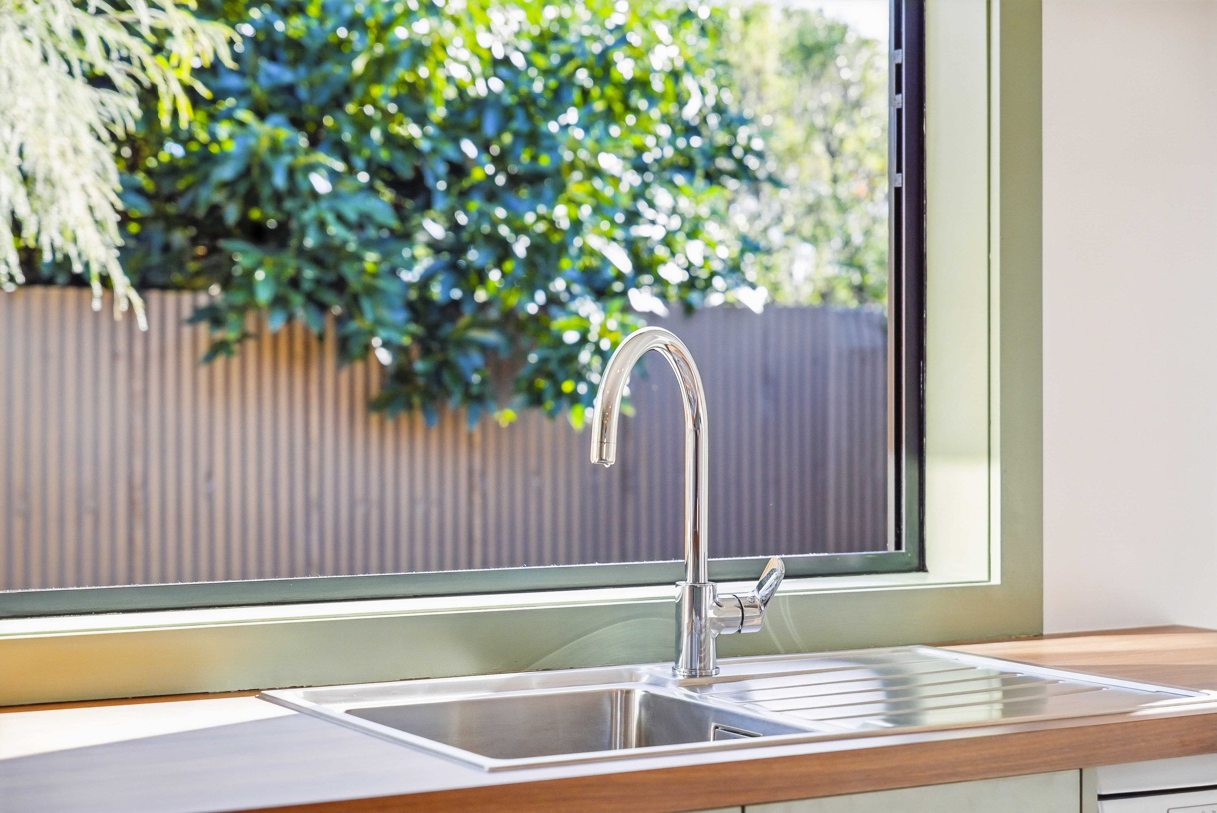 Kitchen sink with a silver faucet viewed through a green-framed window, with trees and a wooden fence outside.