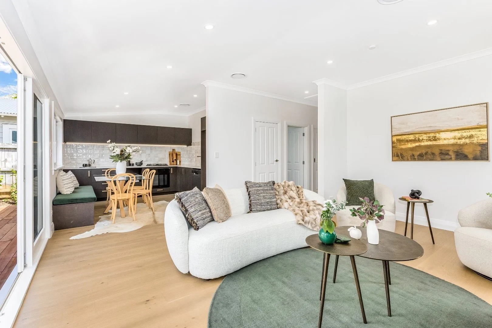 Bright open-concept living room and kitchen with white walls, beige flooring, white and green furniture, and decorative plants.