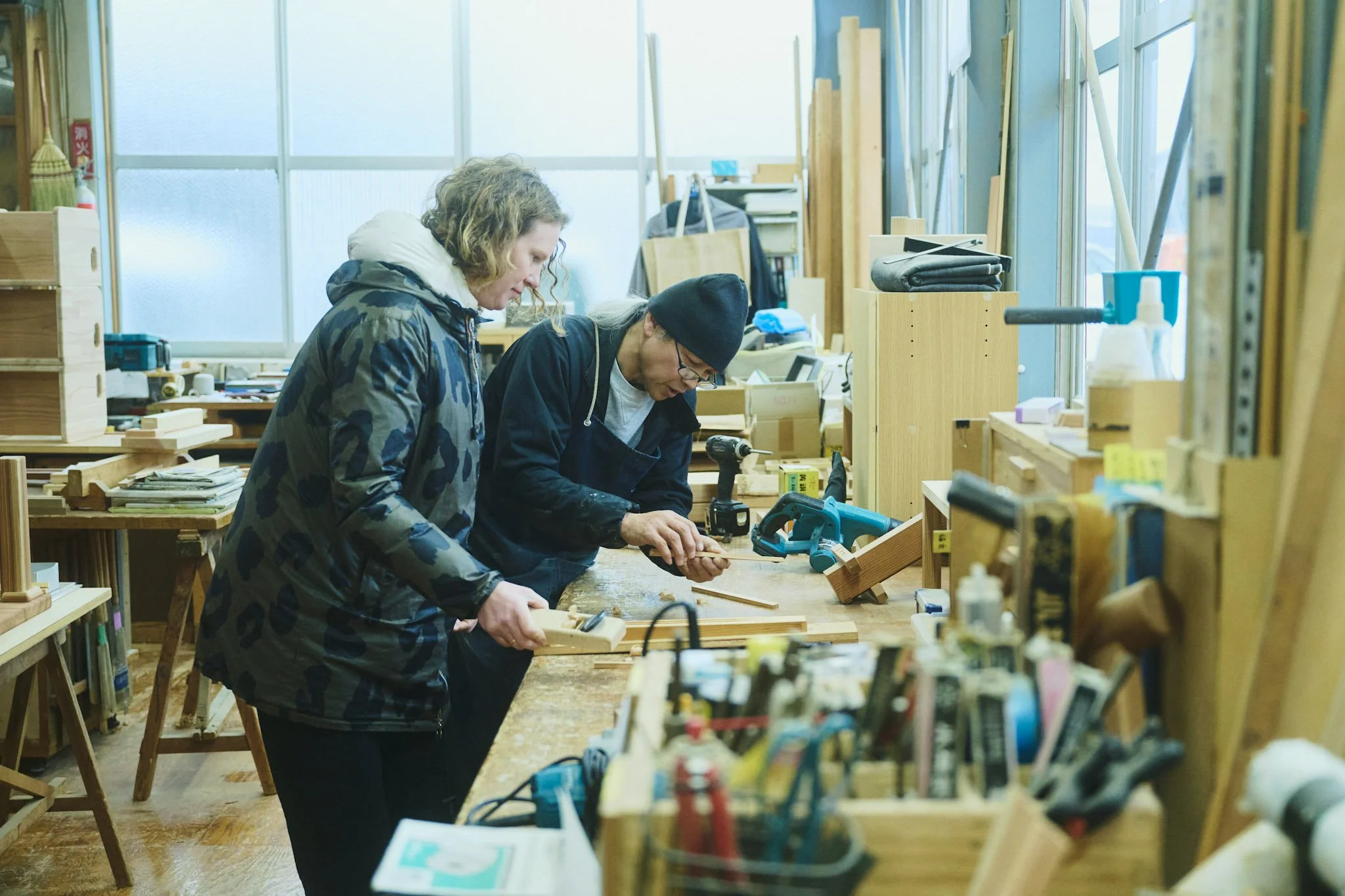 Two people working together on woodworking projects in a workshop, surrounded by tools and wooden pieces.