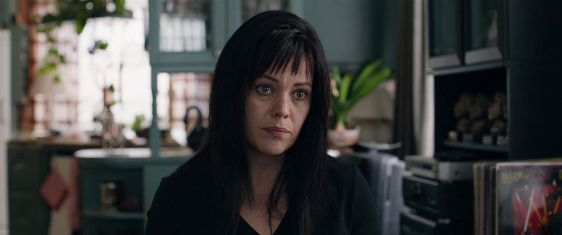 Close-up of a woman with dark hair and pale skin, looking slightly to her right with a serious expression. She is in a room with a window, plants, and shelves filled with objects, possibly a kitchen or living space.