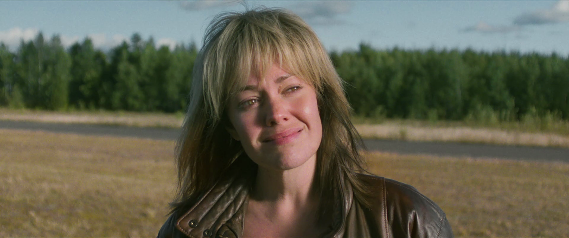 Woman with short blonde and brown hair, wearing a brown leather jacket, standing outdoors in a rural area with fields and trees in the background.