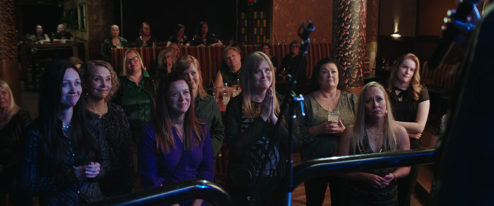 A group of women attending a live performance at a bar or small venue, standing and sitting near the stage, some with drinks, watching attentively with expressions of enjoyment and anticipation.