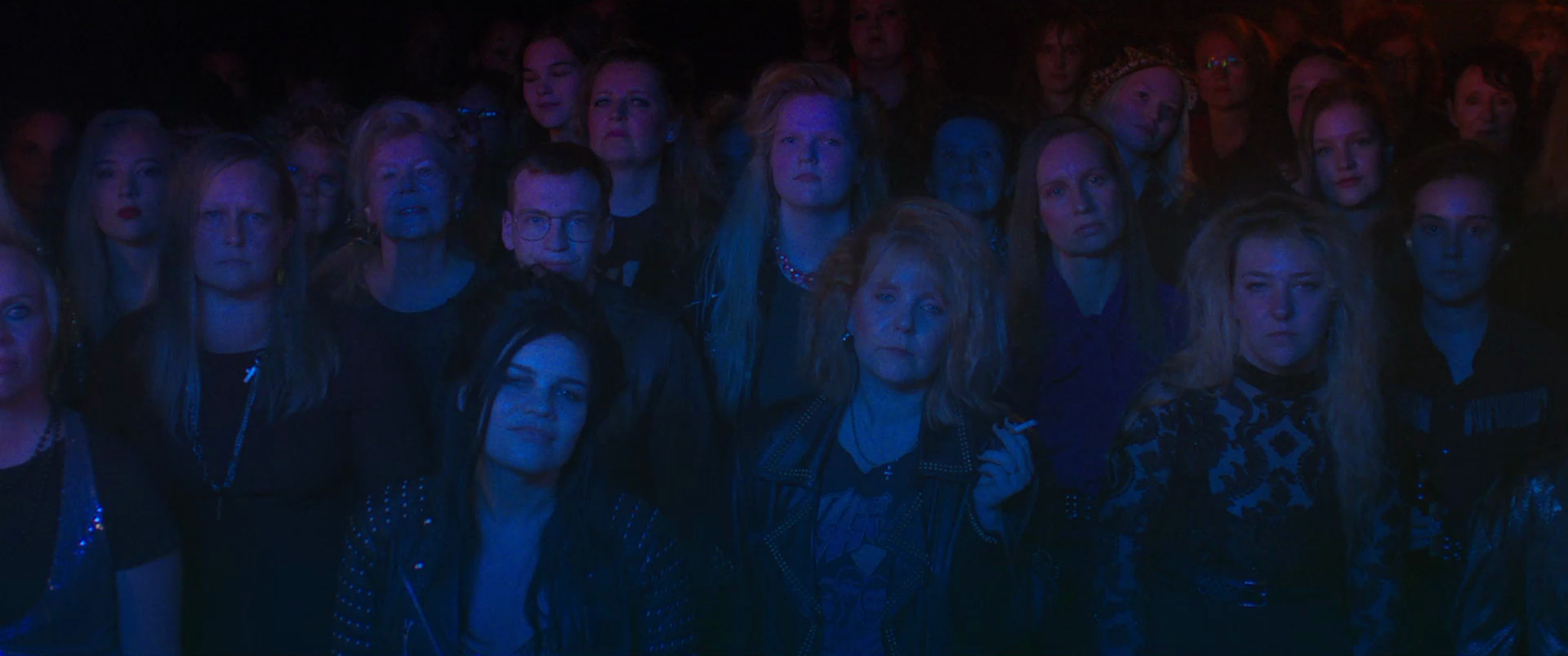 A group of diverse people with serious expressions standing close together in a dark, possibly concert or theater setting with dim blue and red lighting.
