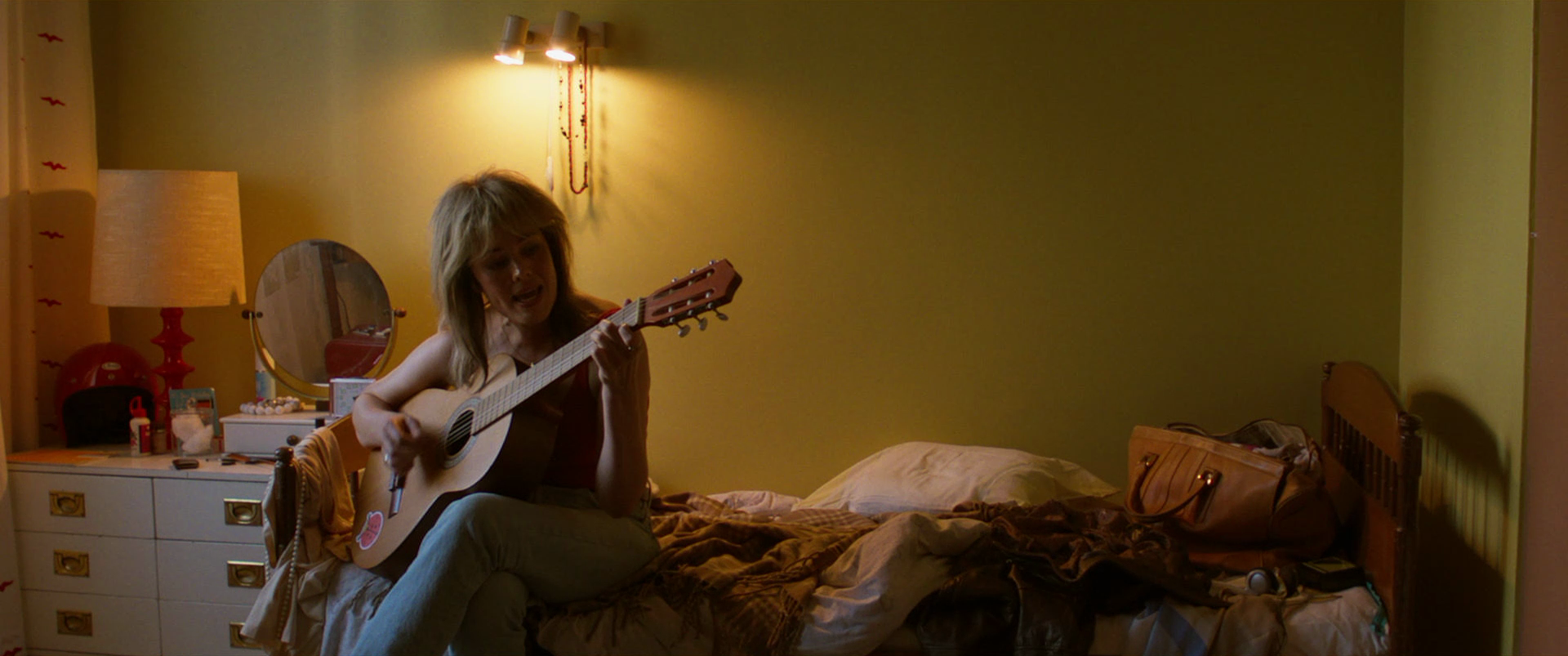 A woman with shoulder-length blonde hair sitting on a bed, playing an acoustic guitar in a dimly lit bedroom with a yellow wall. The bed has a brown and beige blanket, and a dresser with various items and a lamp is visible nearby.