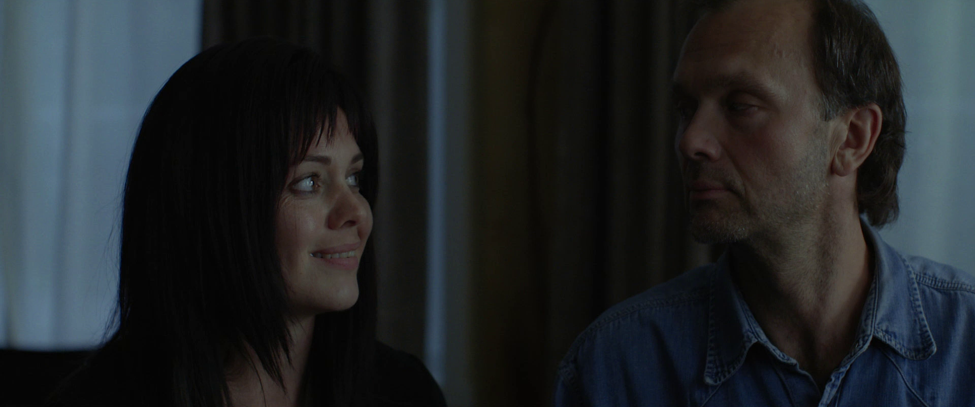 A woman with dark hair smiling and looking at a man with short hair, who is looking back at her, in a dimly lit room.