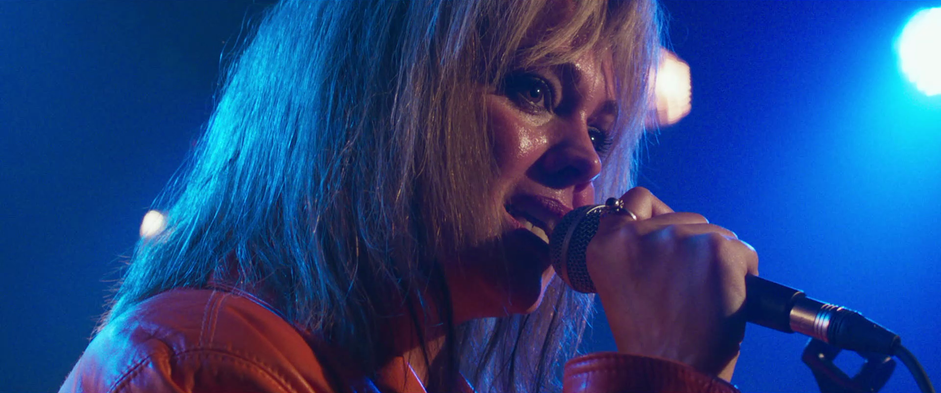 Close-up of a woman with blonde hair singing into a microphone on stage, illuminated by colorful stage lights.