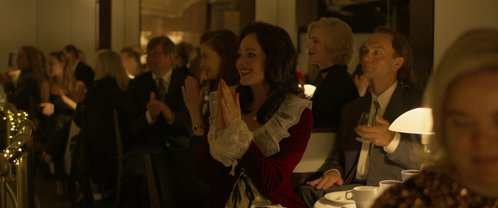 A group of people dressed in formal attire sitting at a table, watching a performance or event with smiles and attentive expressions in a warmly lit room.