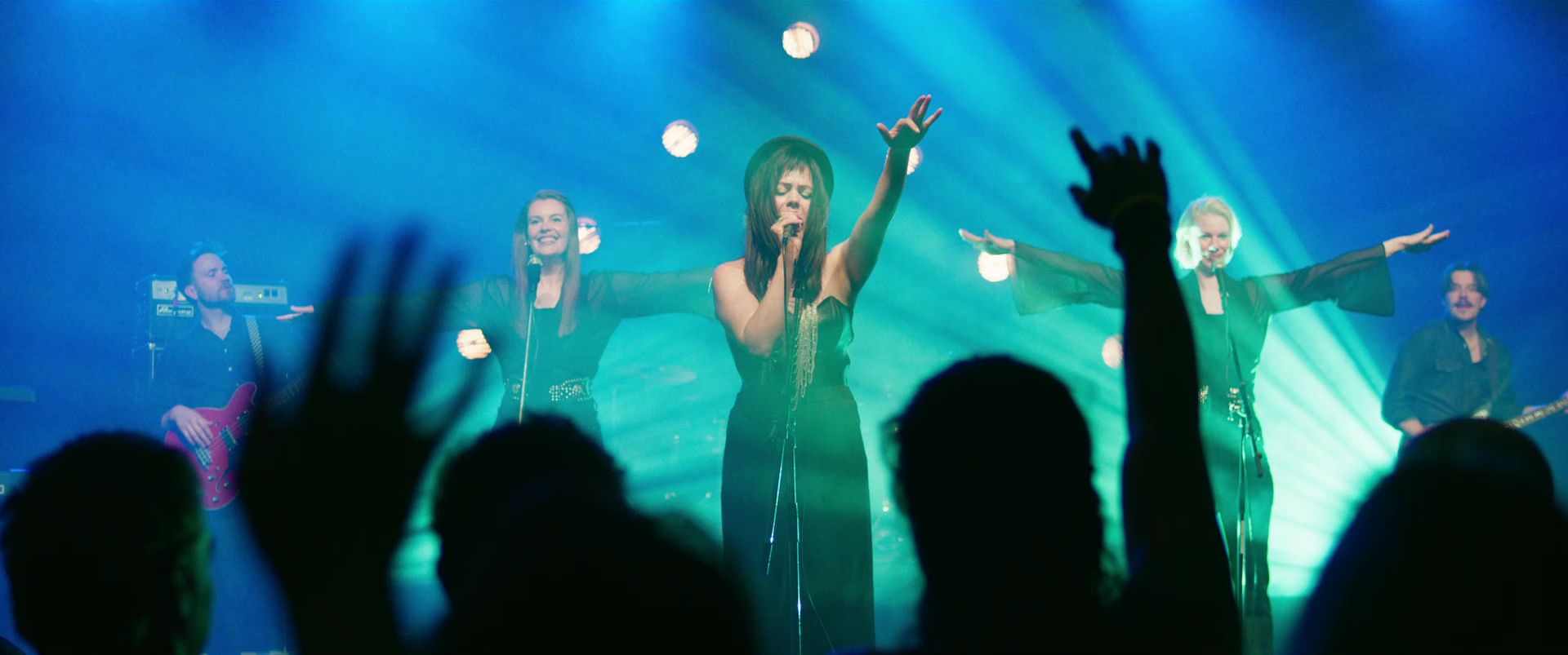 Female singer performing on stage with backup singers and musicians in the background, audience raising their hands.