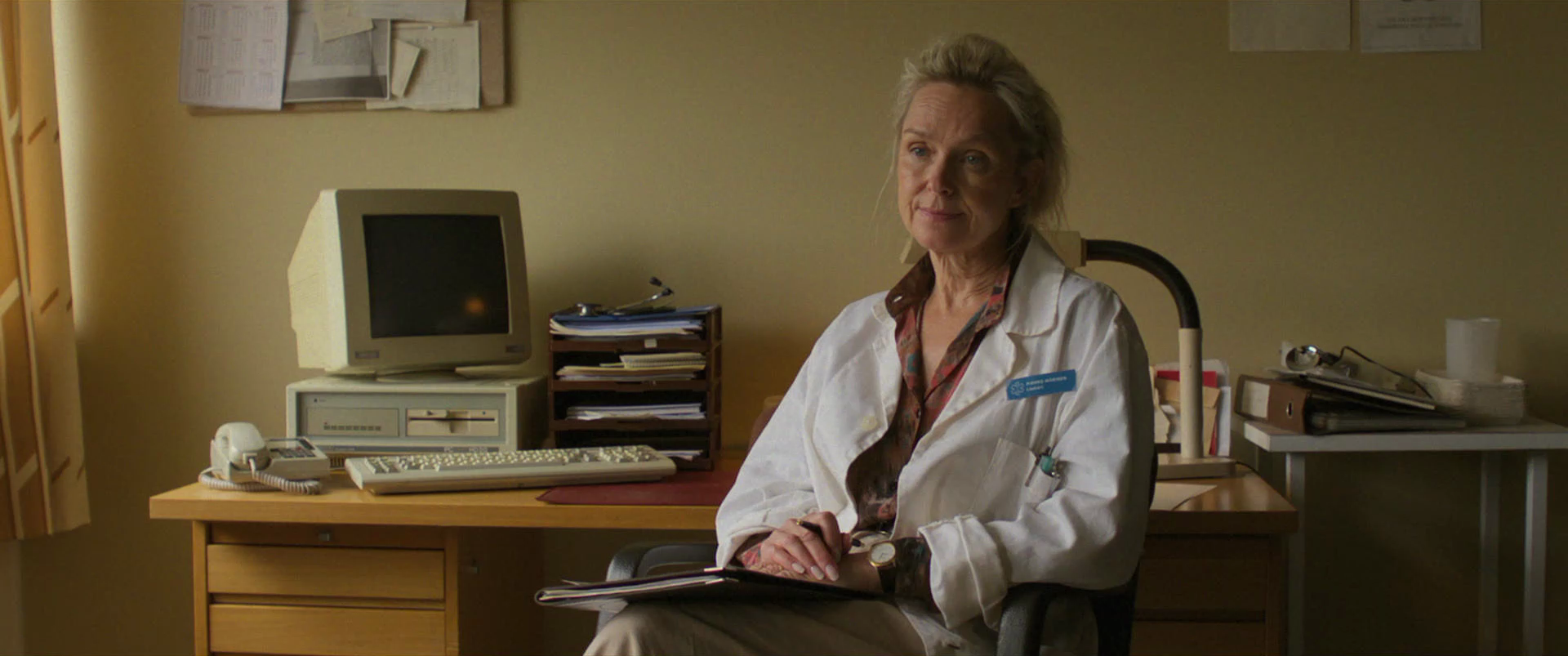 A female doctor sitting at her desk in an office, wearing a white coat with a stethoscope around her neck, holding a clipboard.