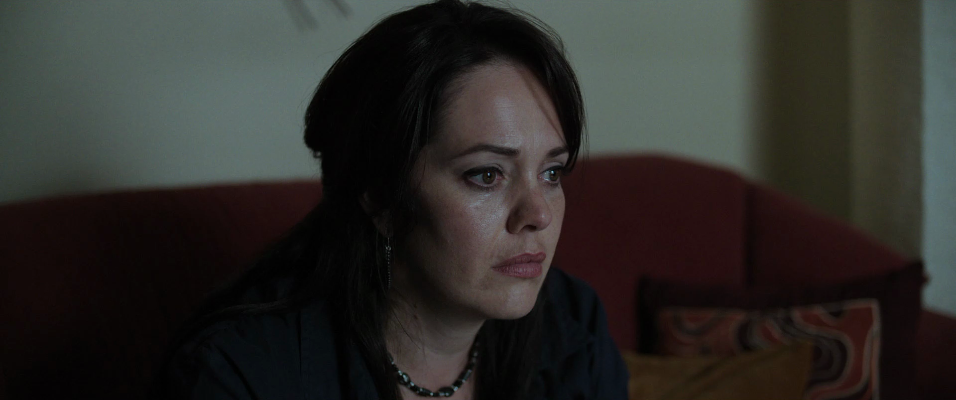 A woman with dark hair and makeup sitting on a red couch, looking contemplative, with a beige wall in the background.