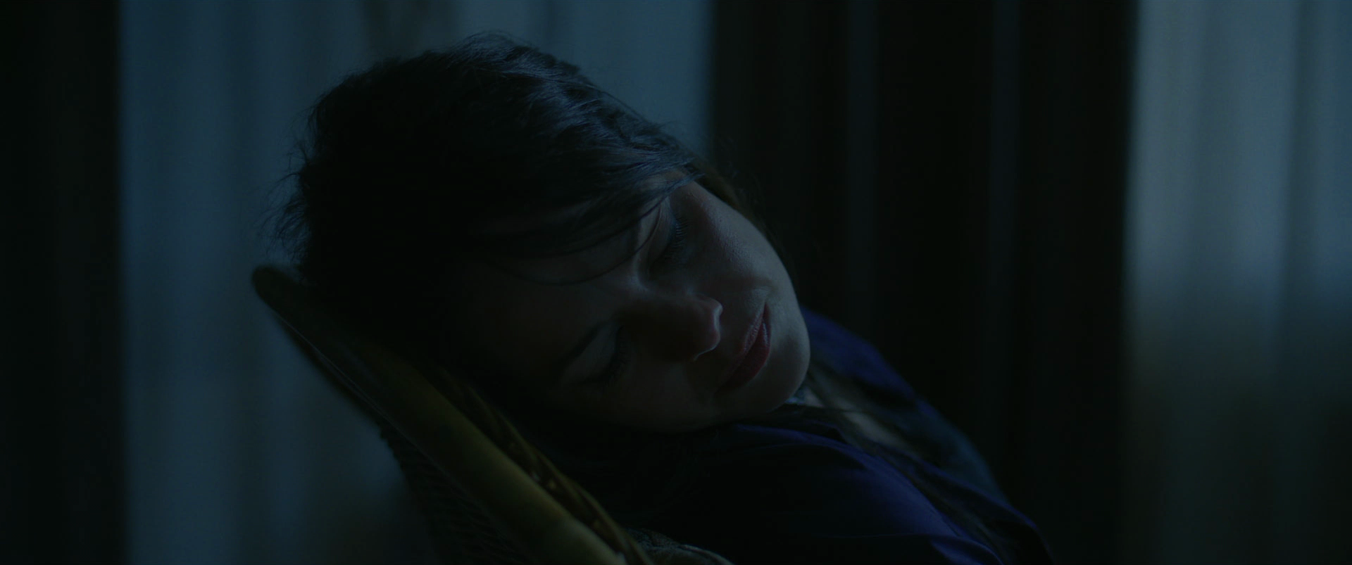 A woman with dark hair and closed eyes resting her head on a yellow pillow, in a dimly lit room.